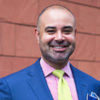 Headshot of a person wearing a bright blue suit, pink shirt, and yellow tie, smiling in front of a brick wall.