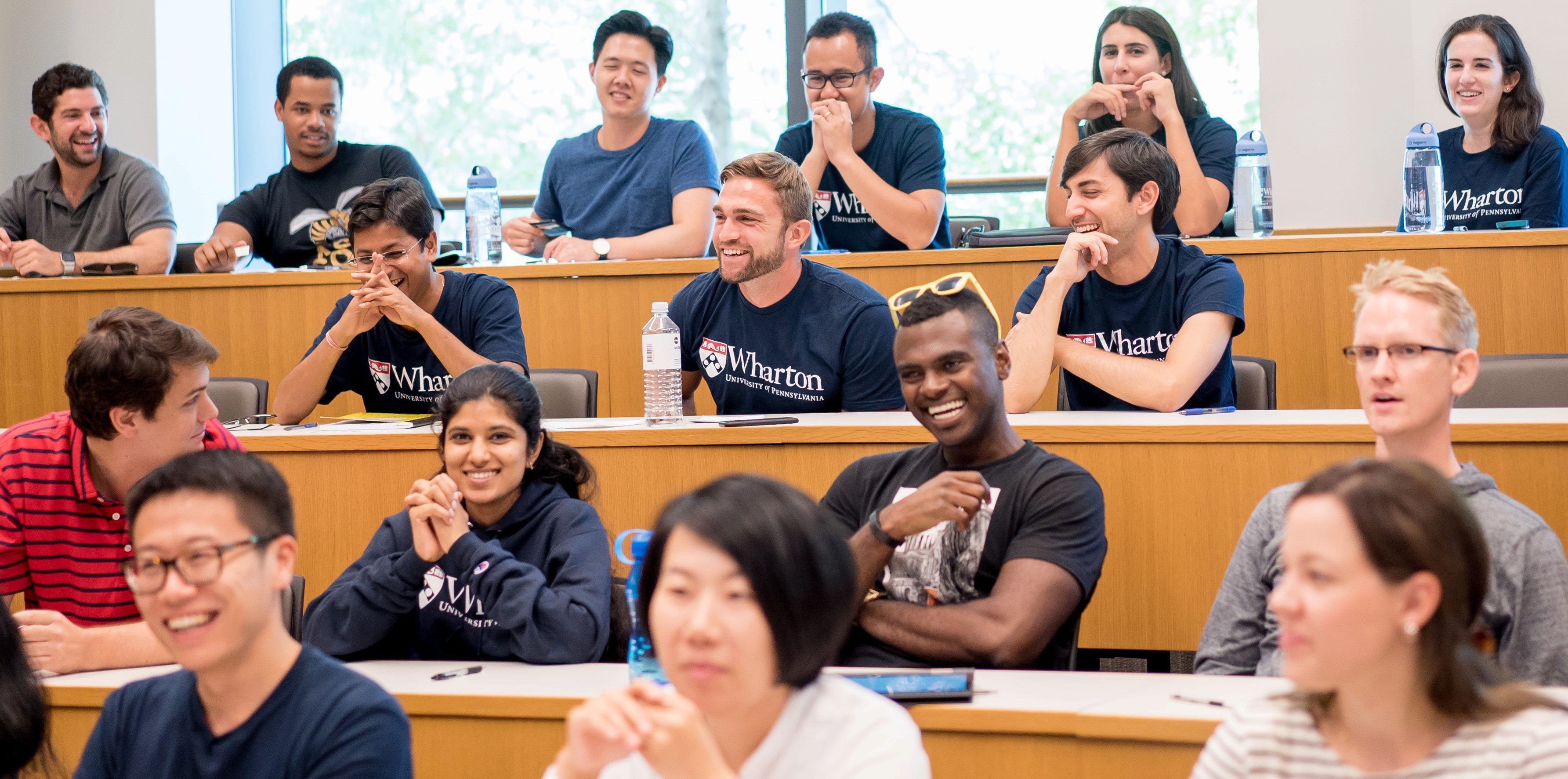 A diverse group of people sitting in a classroom setting, smiling and engaged. Some are wearing shirts with Wharton University of Pennsylvania logos.