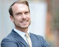 Headshot of a person wearing a suit and tie, smiling, with an outdoor background.