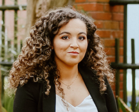 Headshot of a person with curly hair wearing a black blazer, standing outdoors in front of a brick wall and metal fencing.