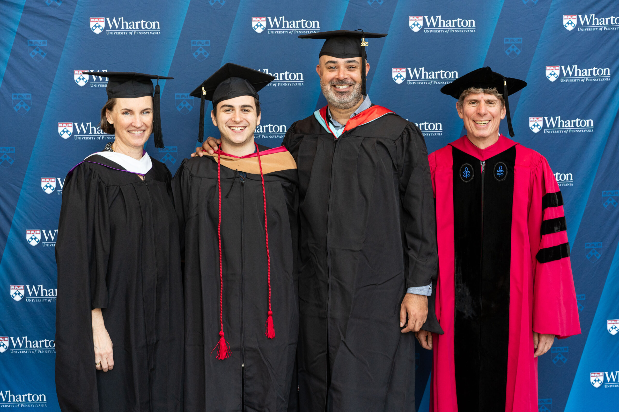 A group of people dressed in academic regalia, including caps and gowns, stand in front of a backdrop featuring the Wharton School logo, indicating a graduation ceremony.