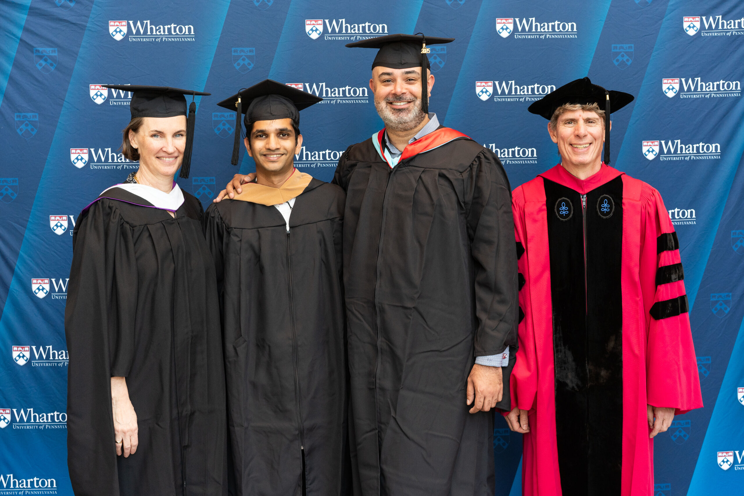 A group in academic regalia stands in front of a Wharton School backdrop, likely at a graduation event.