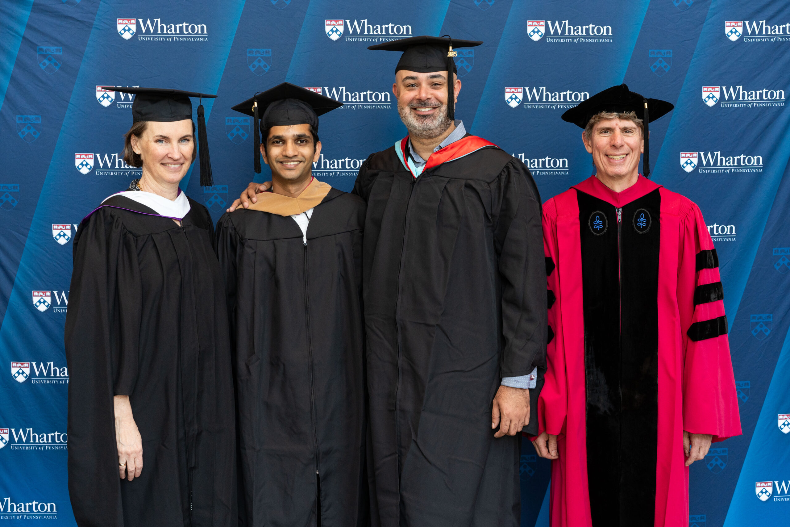 A small group dressed in graduation caps and gowns poses in front of a Wharton School backdrop.