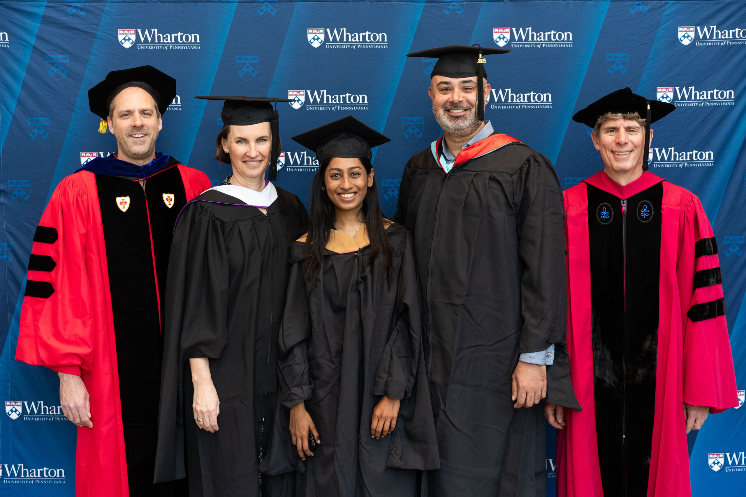 A group of people in graduation caps and gowns, standing in front of a Wharton School backdrop. Some are in ceremonial robes, indicating a graduation or academic event.