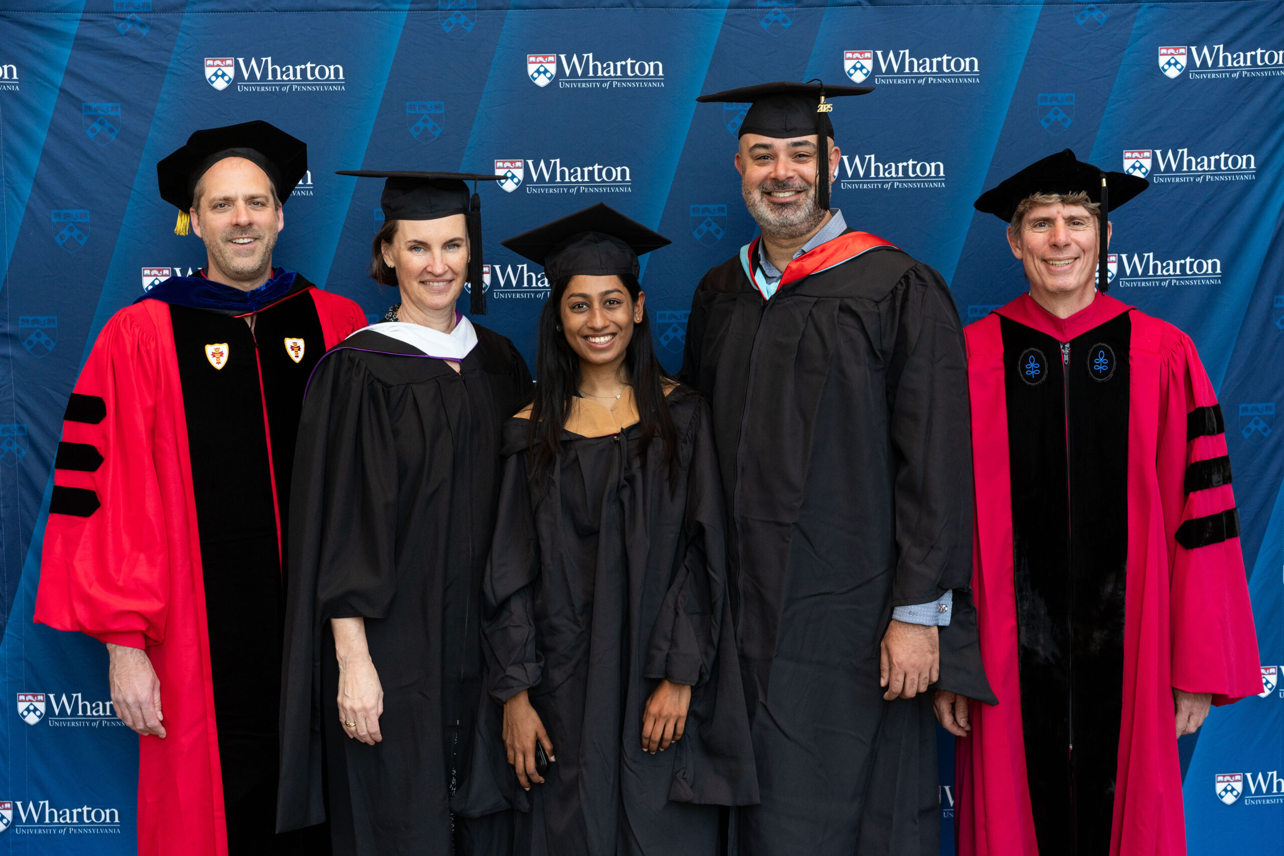 A group of people in academic regalia standing in front of a Wharton School of the University of Pennsylvania backdrop.