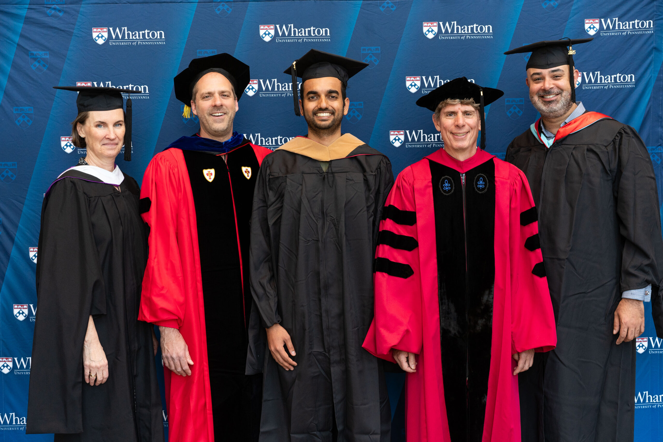 A group of people in academic regalia standing in front of a Wharton School banner. Some are wearing red robes, and others are in black graduation gowns and caps, suggesting a graduation ceremony.