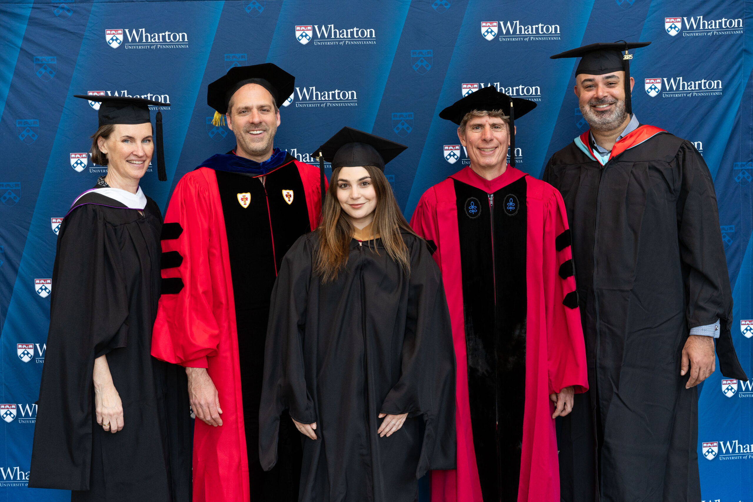 A small group of individuals in academic regalia, including caps and gowns, stand in front of a Wharton School backdrop, appearing to celebrate a graduation event.
