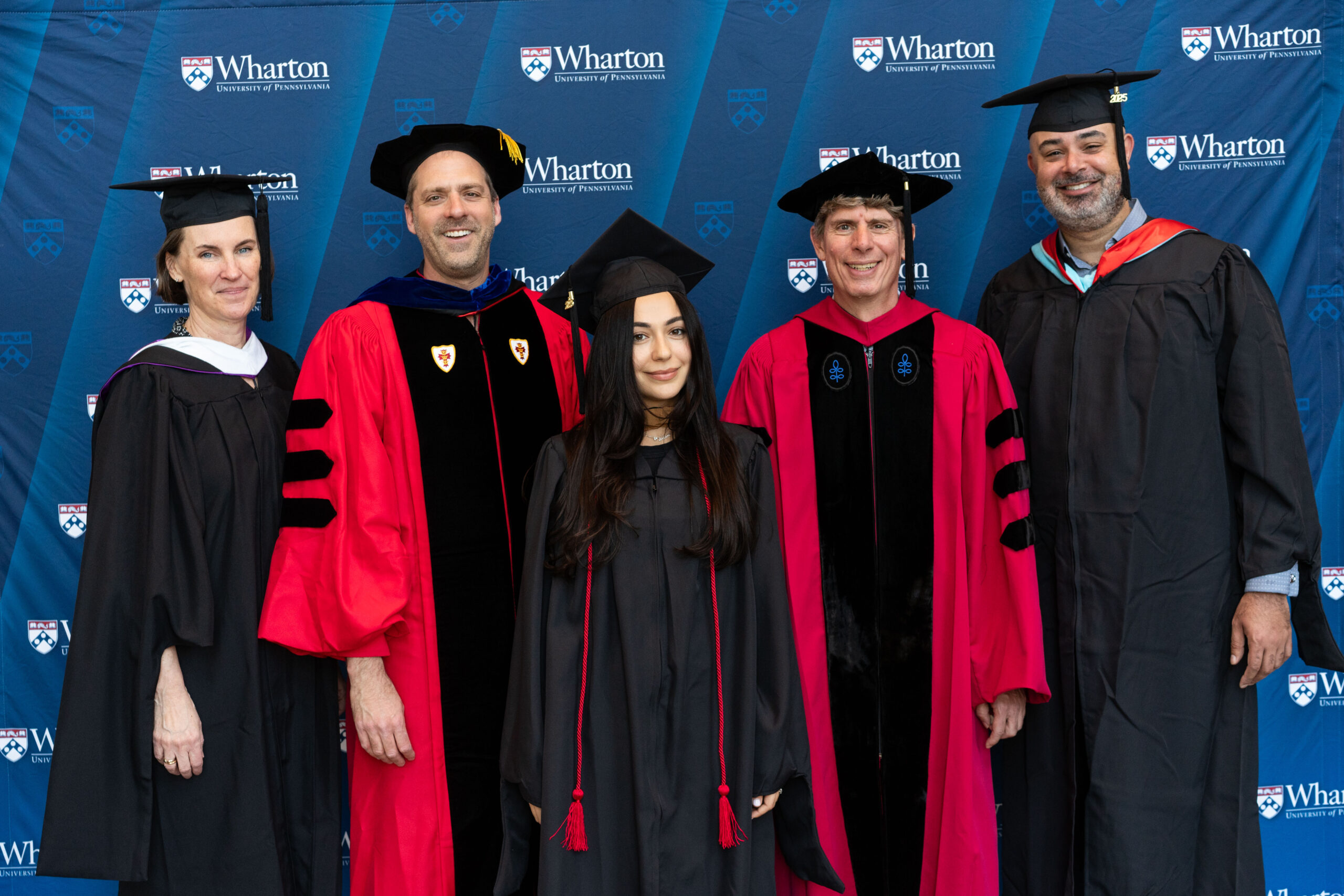 A group of individuals in graduation attire posed in front of a Wharton School backdrop. Some are in black gowns, others in red academic regalia.
