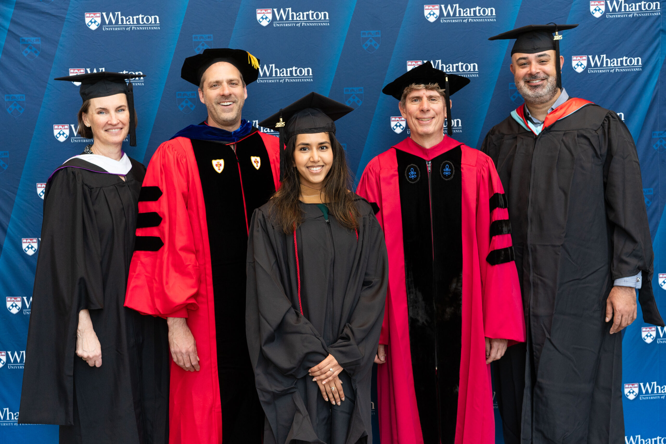 A group of people in academic regalia, including robes and caps, stands in front of a backdrop featuring the Wharton School logo, suggesting a graduation event.
