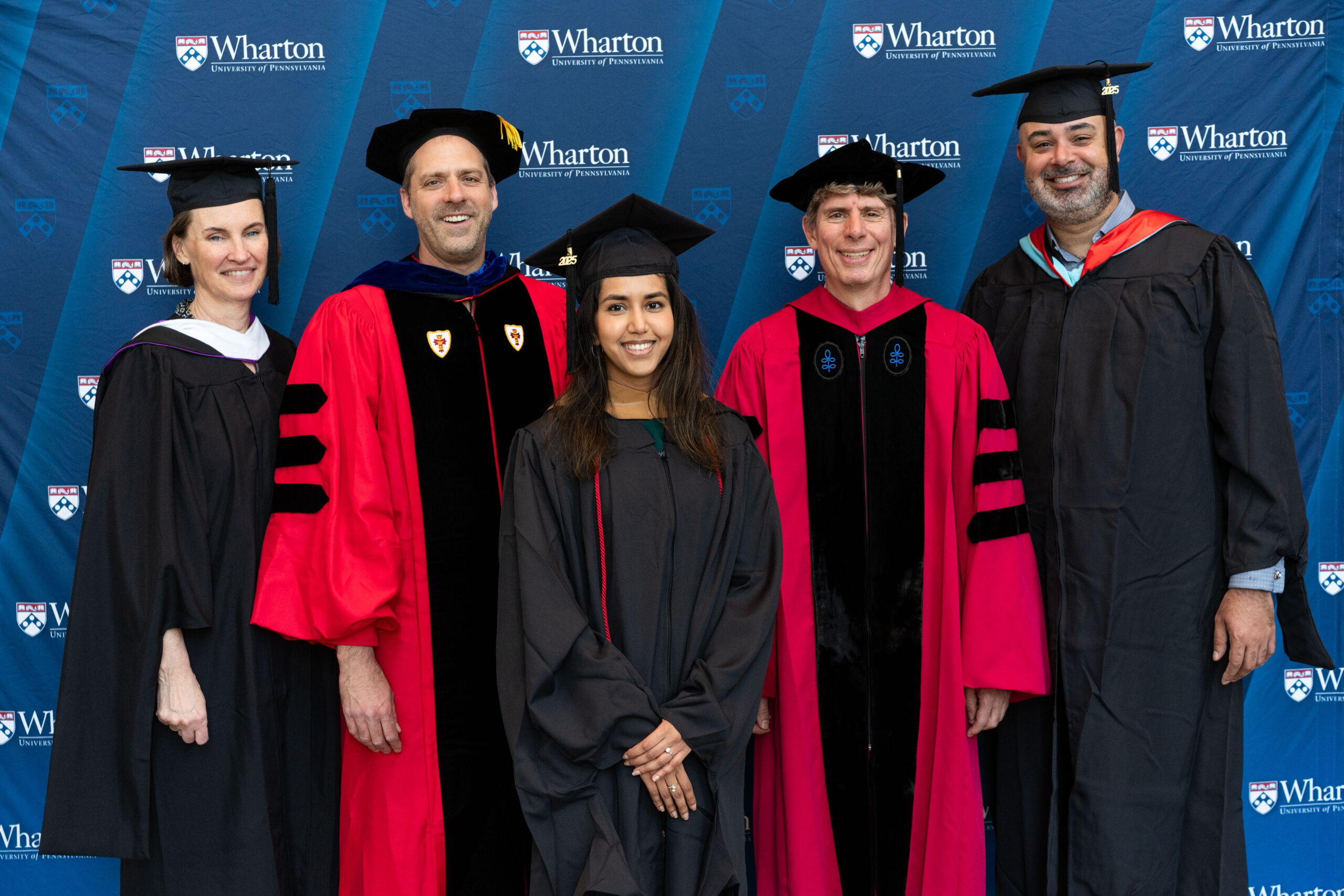 A small group of people in academic regalia, including caps and gowns, posing in front of a Wharton School banner, likely at a graduation ceremony.