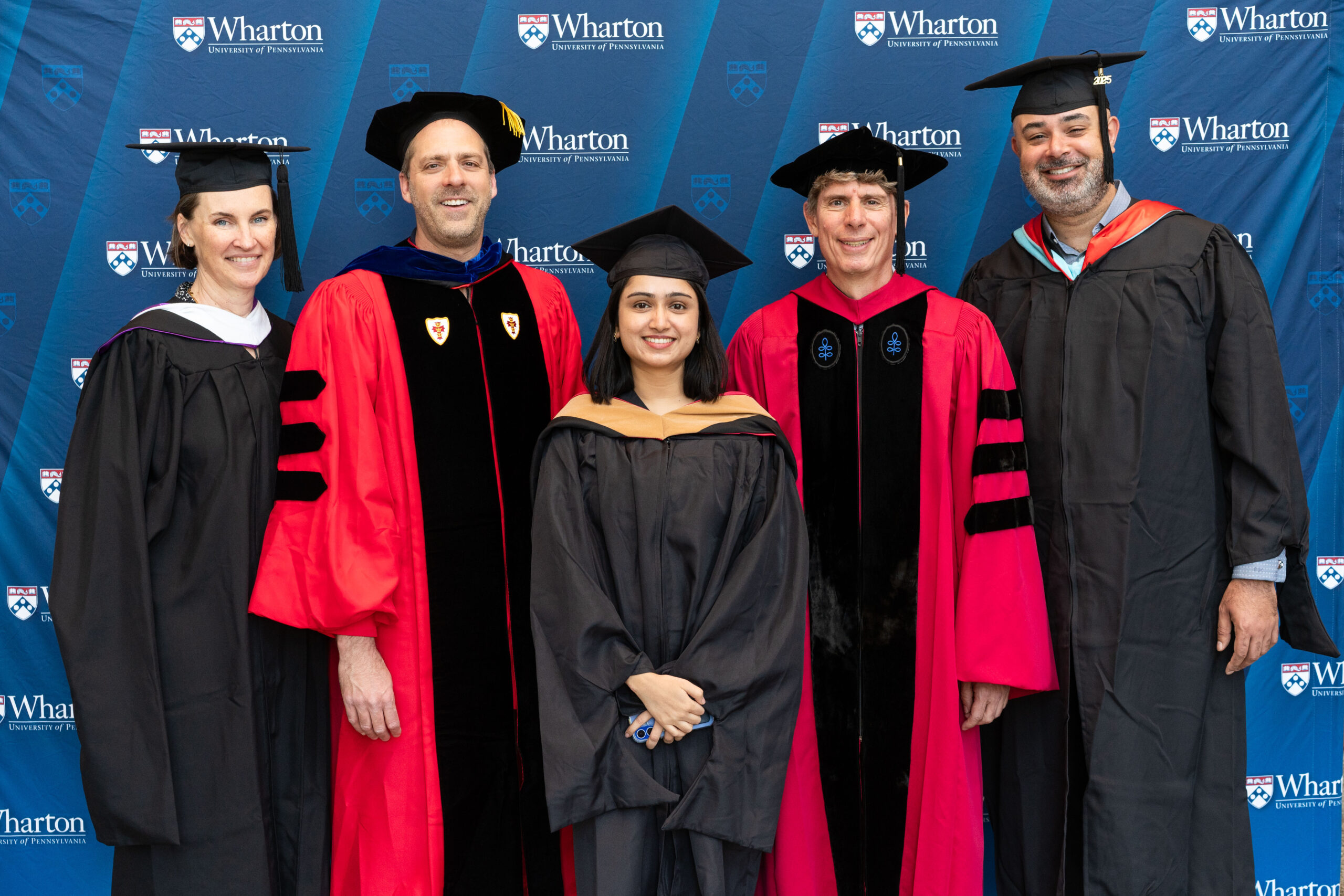 Several individuals in graduation attire, including caps and gowns, stand in front of a Wharton School backdrop, suggesting a graduation event.