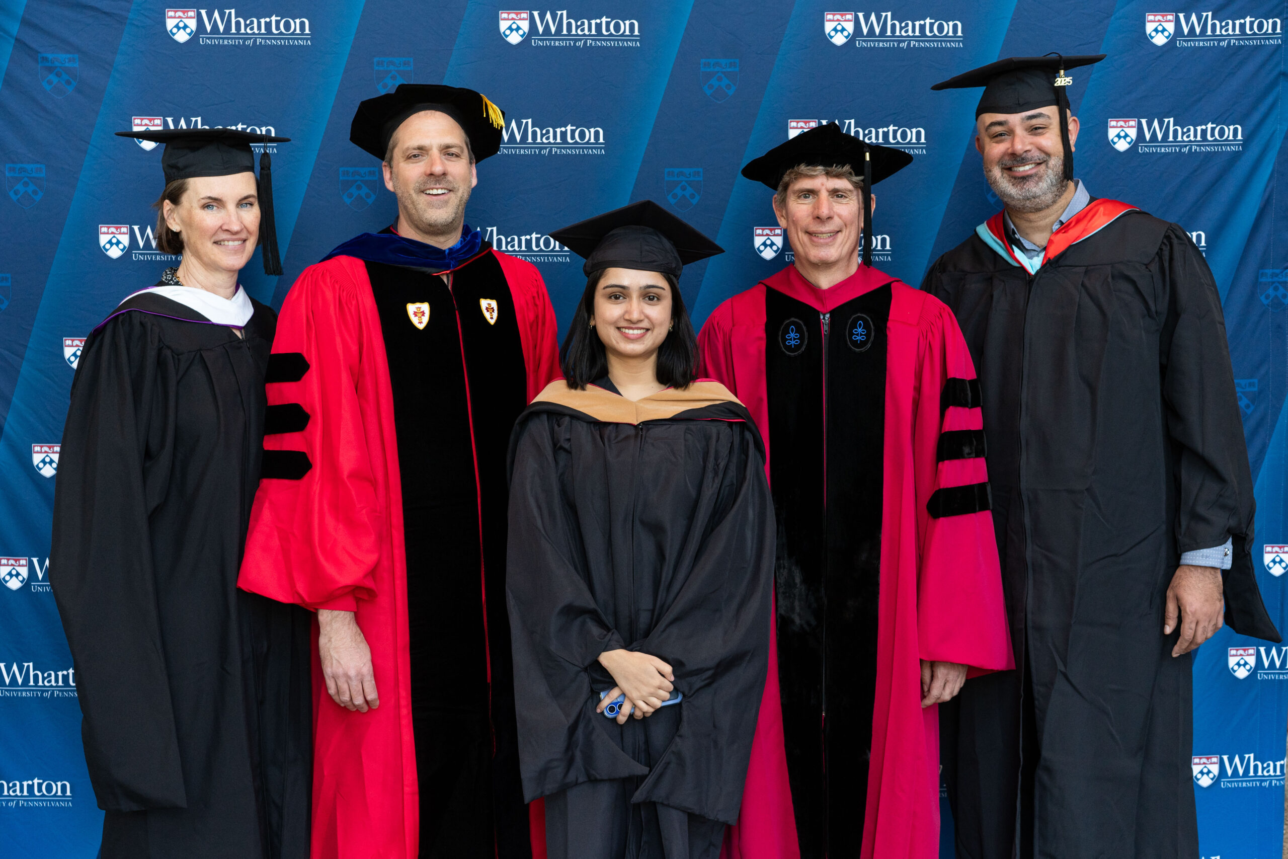 A small group of people wearing academic regalia, including caps and gowns, likely at a graduation event. They are standing in front of a backdrop with the Wharton School logo.