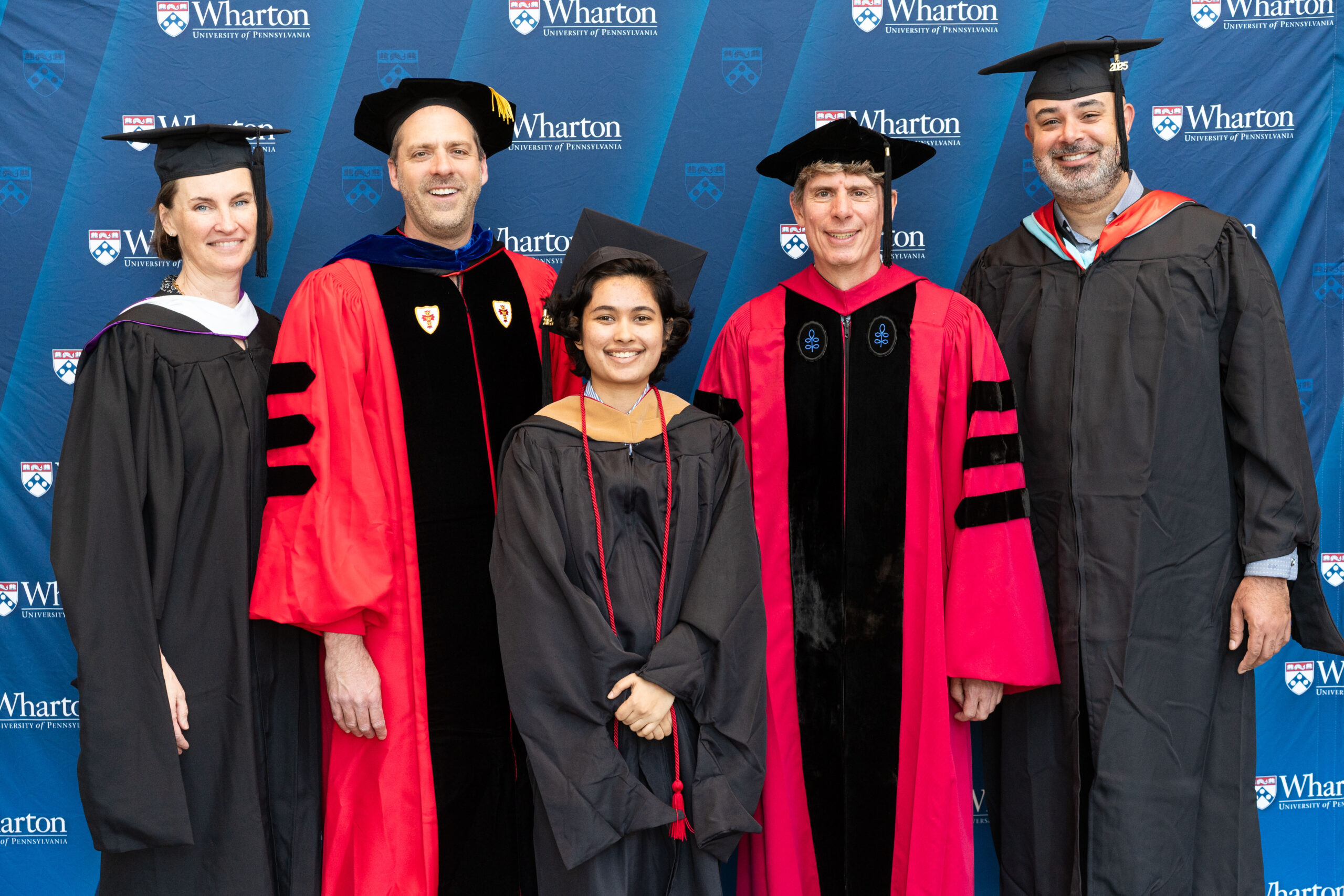 A small group of people in academic regalia, including caps and gowns, standing in front of a Wharton School background. This represents an academic or graduation event.