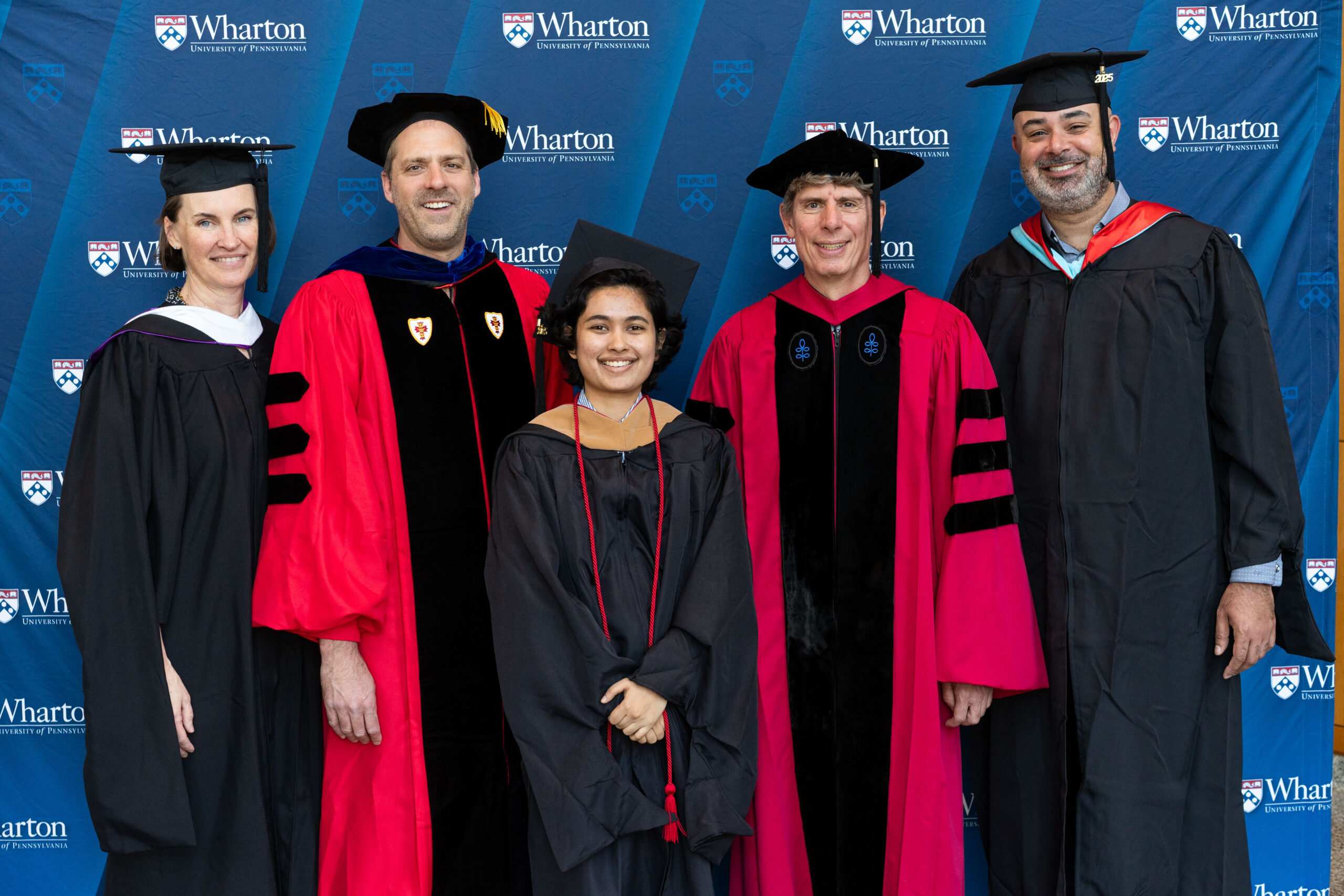 A group of five people in academic robes and caps, likely at a graduation ceremony, standing in front of a Wharton School background.
