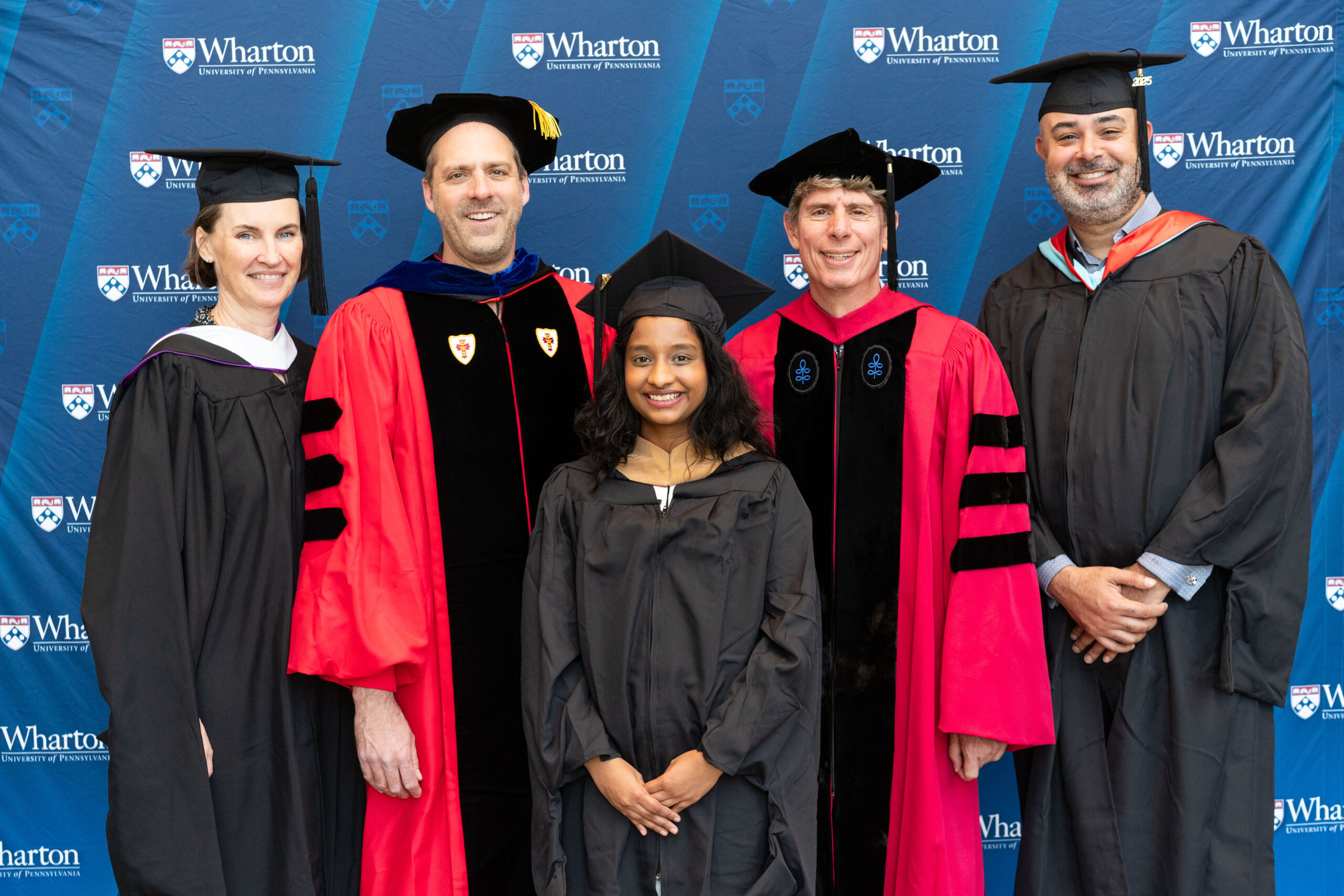 A group of people in graduation attire, including gowns and caps, posing in front of a "Wharton University of Pennsylvania" backdrop. Some are wearing academic regalia with hoods and caps.