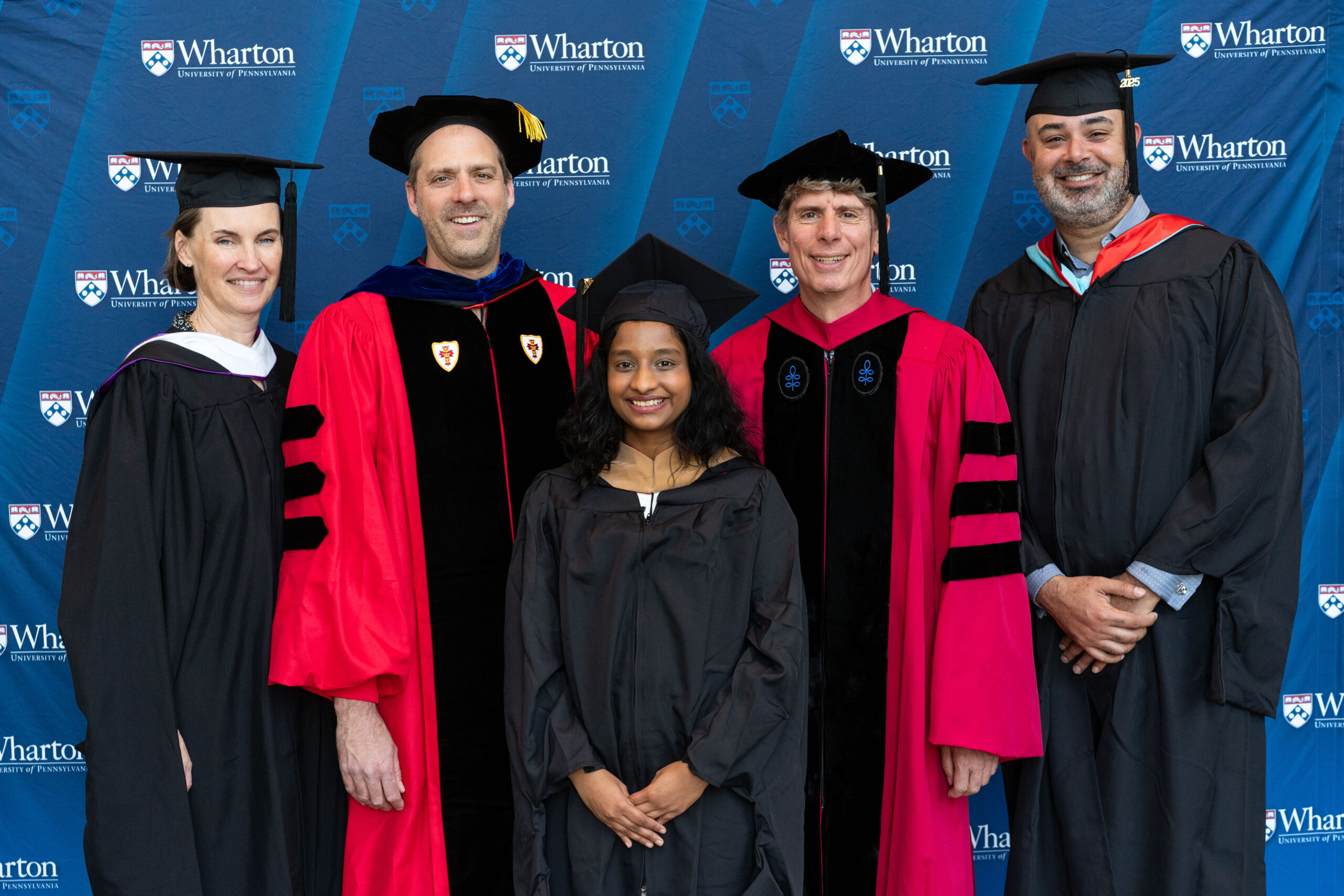 A group of people in academic regalia stand in front of a Wharton School of the University of Pennsylvania backdrop, celebrating a graduation event.