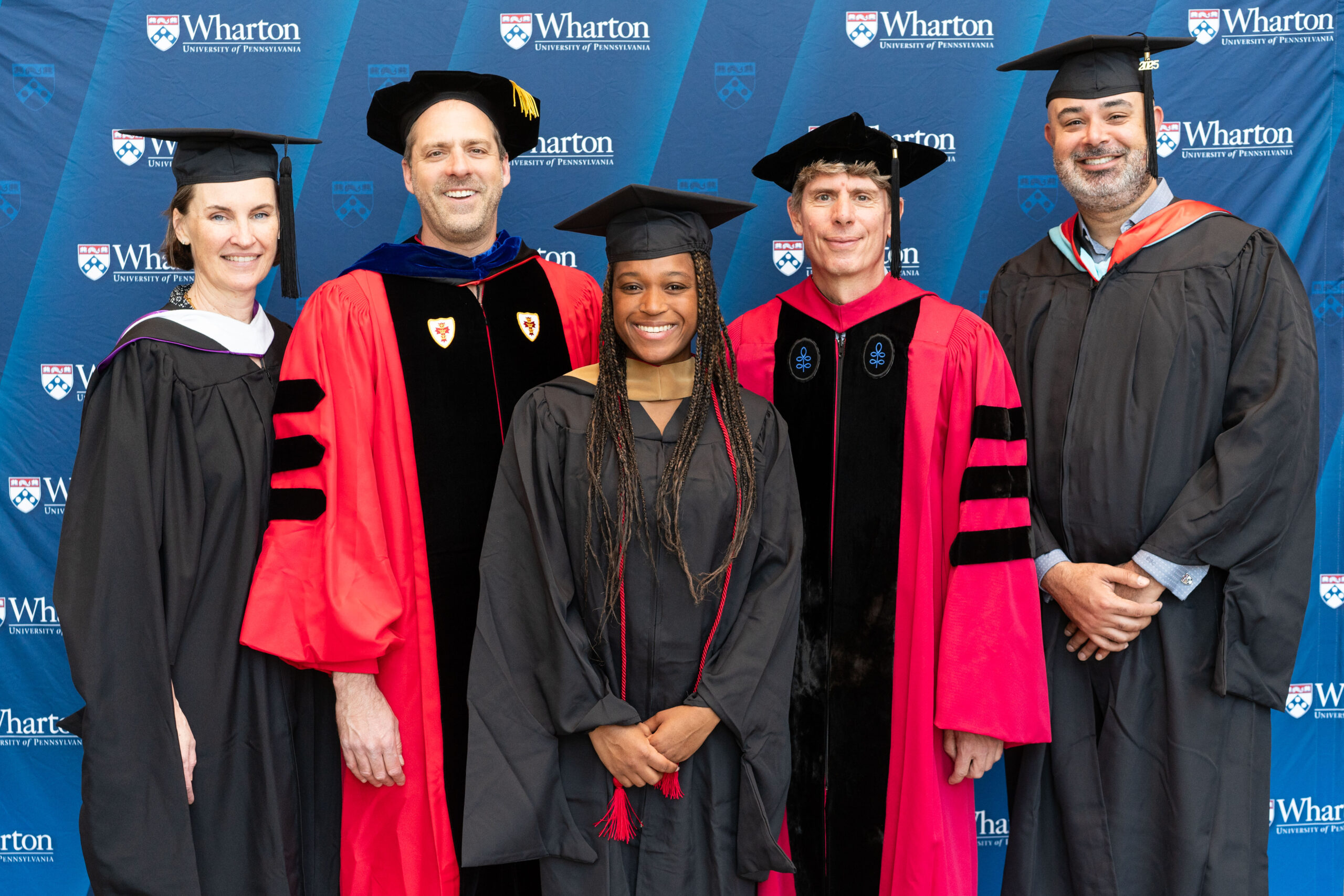A group of people wearing academic regalia, including caps and gowns, posing in front of a backdrop with the Wharton and University of Pennsylvania logos, suggesting a graduation or academic event.