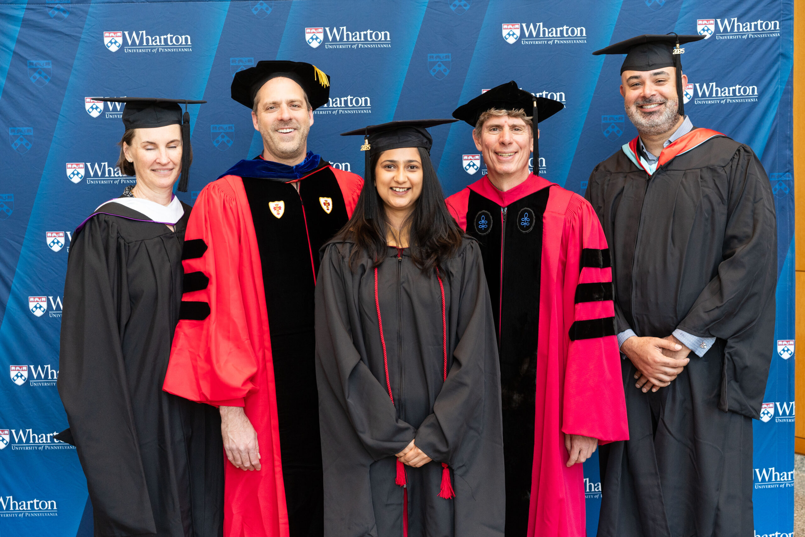 A group of people in academic regalia, including graduation gowns and caps, standing in front of a Wharton School backdrop.
