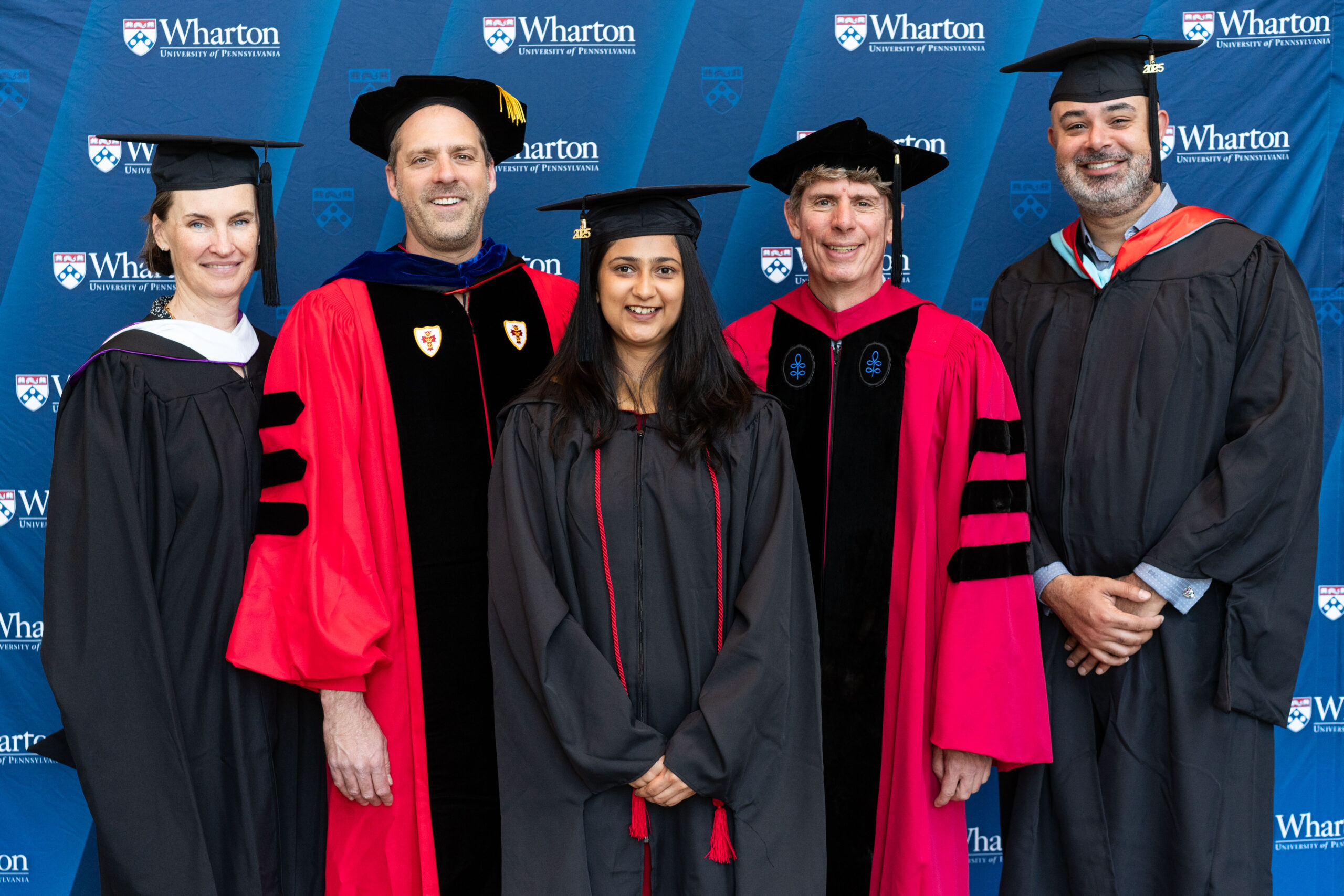 A small group of people in academic regalia, including caps and gowns, standing together at a graduation ceremony in front of a backdrop featuring the Wharton School logo.