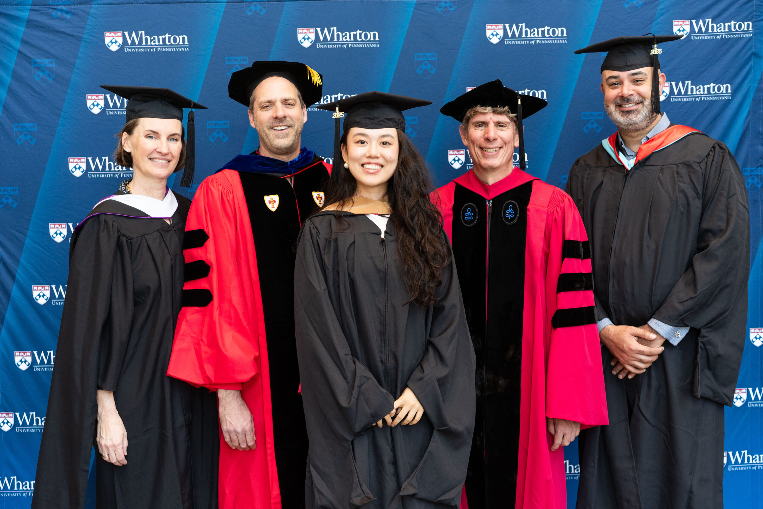 A group of individuals in graduation attire posing in front of a Wharton School backdrop. The scene depicts a celebratory graduation moment.
