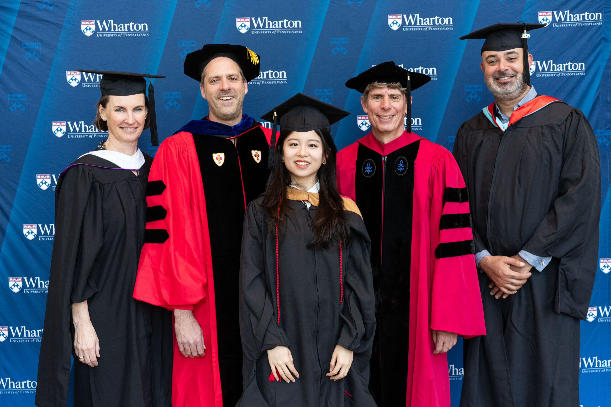 A group of people in academic regalia, including caps and gowns, standing in front of a "Wharton University of Pennsylvania" backdrop.
