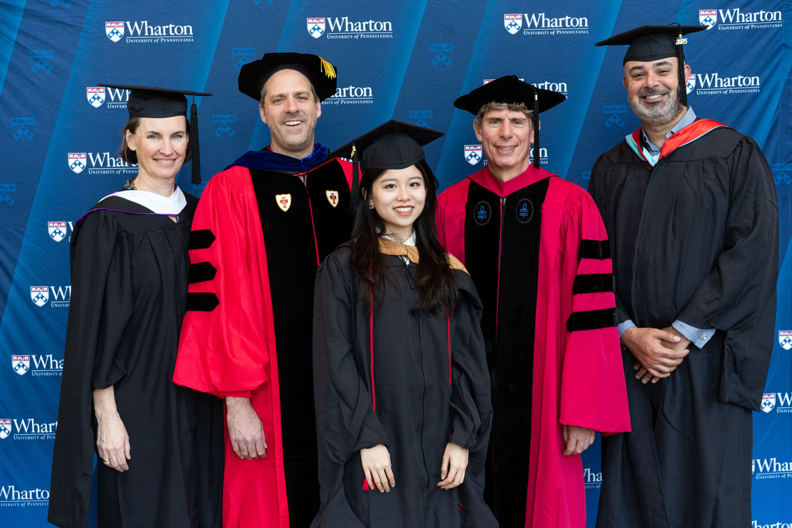 A group of people in academic regalia, including caps and gowns, stand smiling in front of a backdrop with "Wharton University of Pennsylvania" logos, suggesting a graduation or academic event.