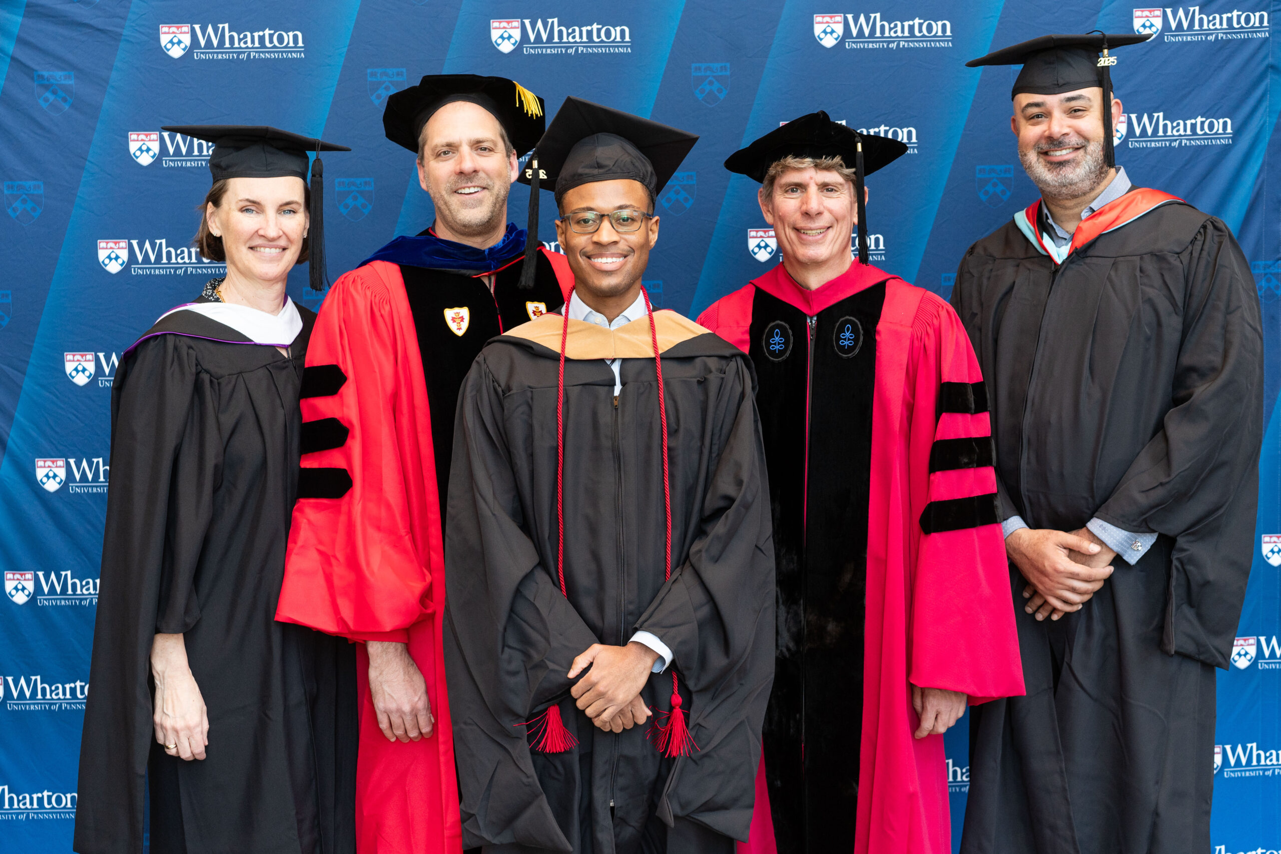 A small group of graduates and faculty members in academic regalia, standing in front of a Wharton School of the University of Pennsylvania backdrop, smiling for a photo.