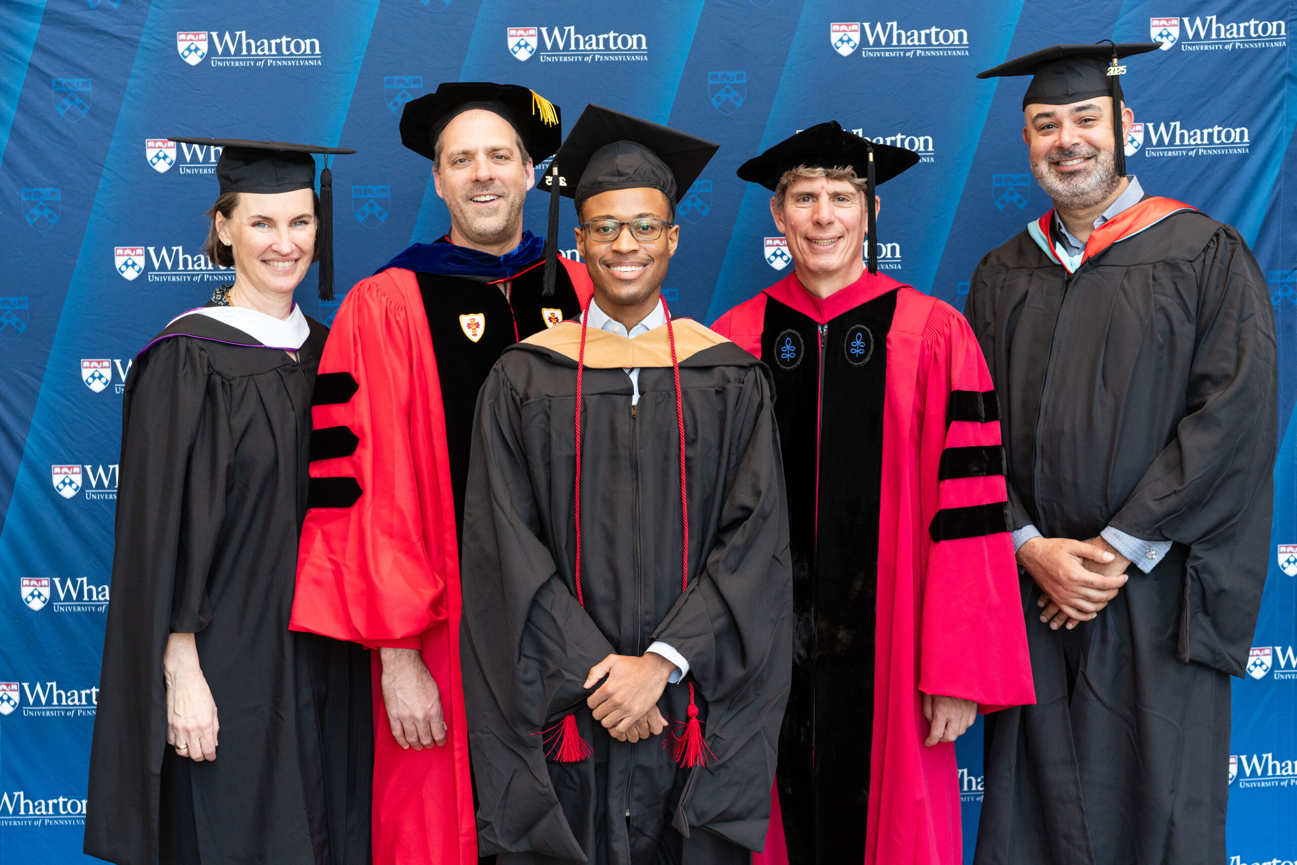 A group of people wearing graduation gowns and caps standing in front of a Wharton School of the University of Pennsylvania backdrop.