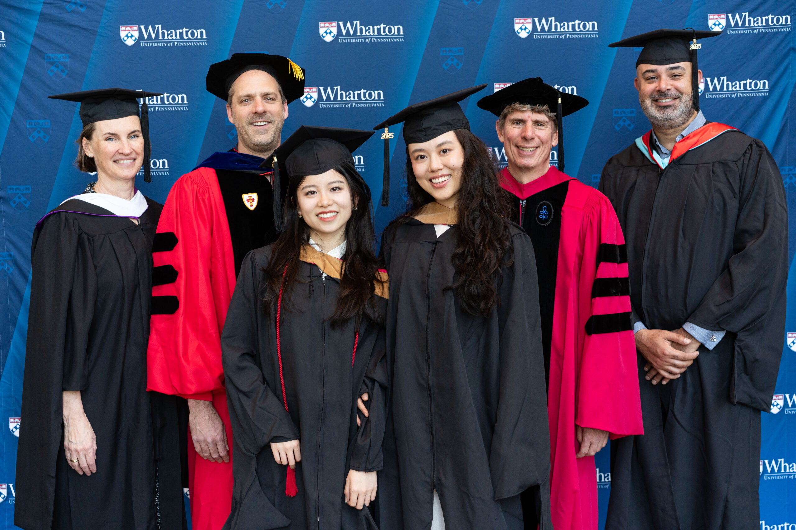 Graduates in caps and gowns pose with faculty members in academic regalia against a backdrop with the Wharton School's logo.