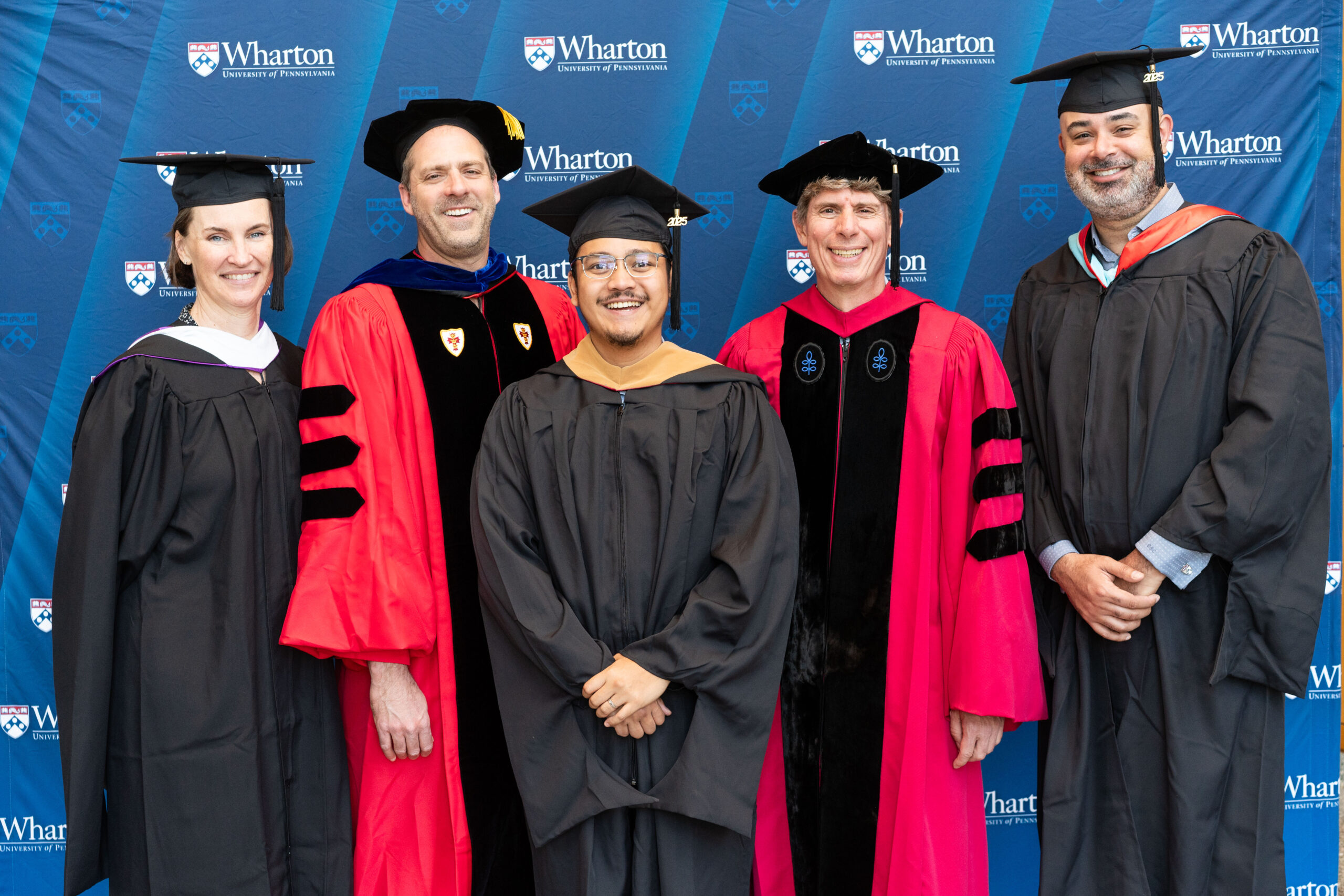A small group of people in graduation attire, including caps and gowns, standing in front of a "Wharton University of Pennsylvania" backdrop.