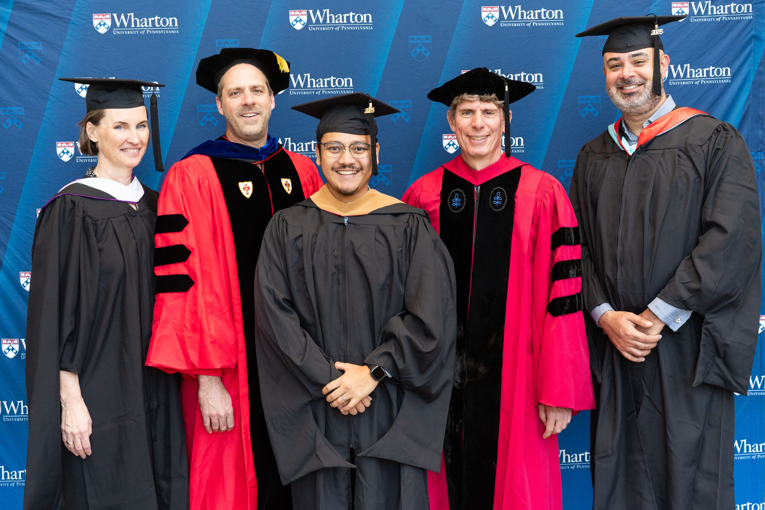 A small group wearing academic gowns and caps, standing in front of a backdrop featuring the Wharton School logo.