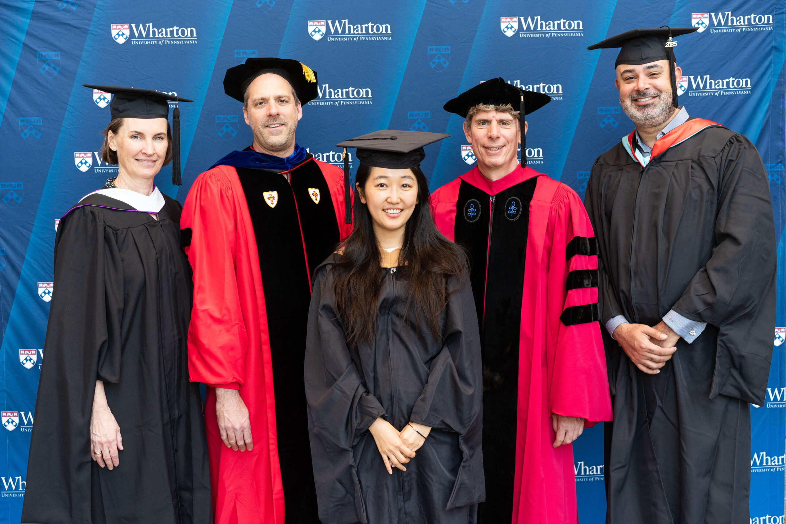 A group of individuals in academic regalia, including caps and gowns, posing in front of a Wharton School of the University of Pennsylvania backdrop.