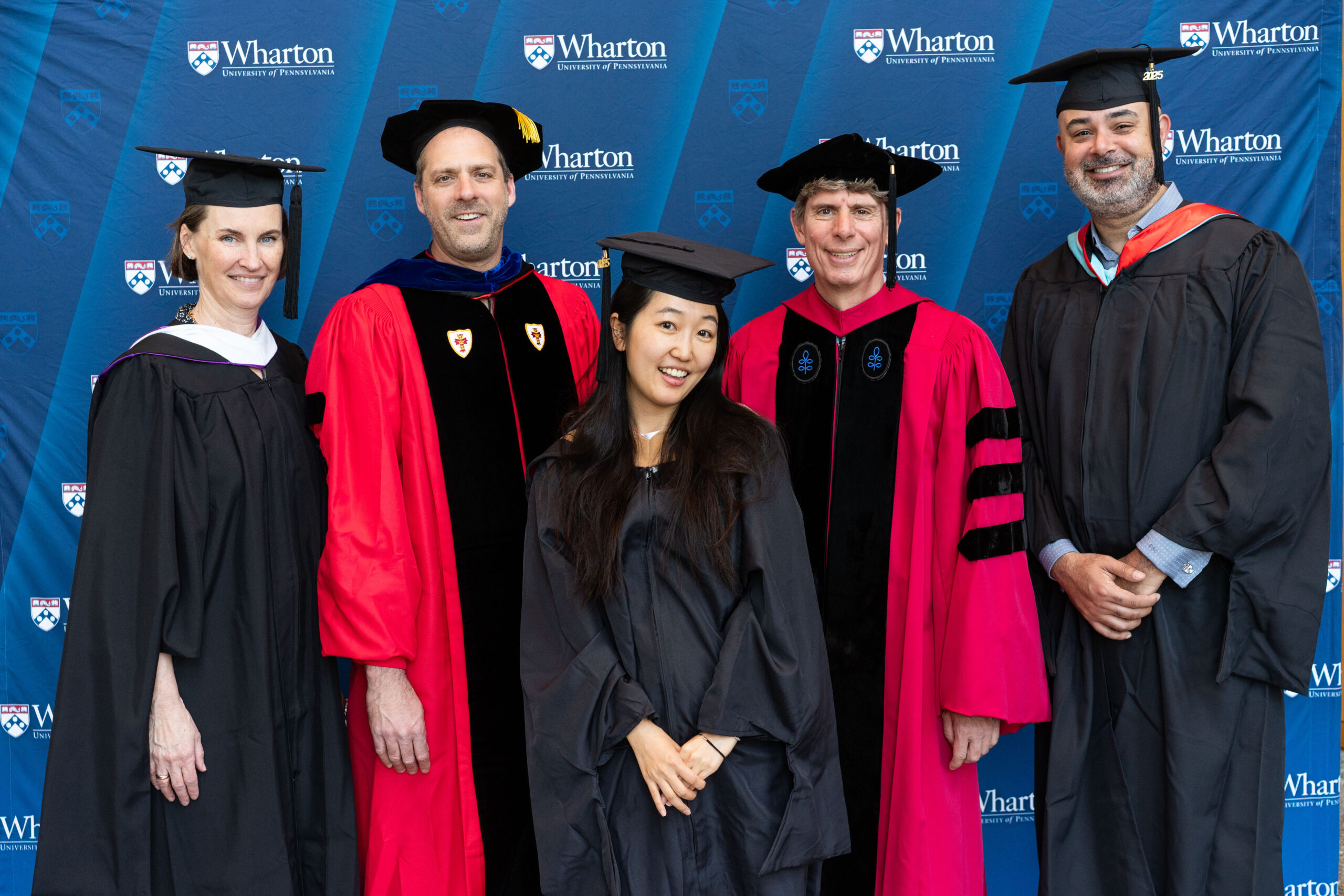 A small group of people in graduation attire posing for a photo in front of a Wharton School backdrop. Some are in red academic robes, others in black graduation gowns and caps.