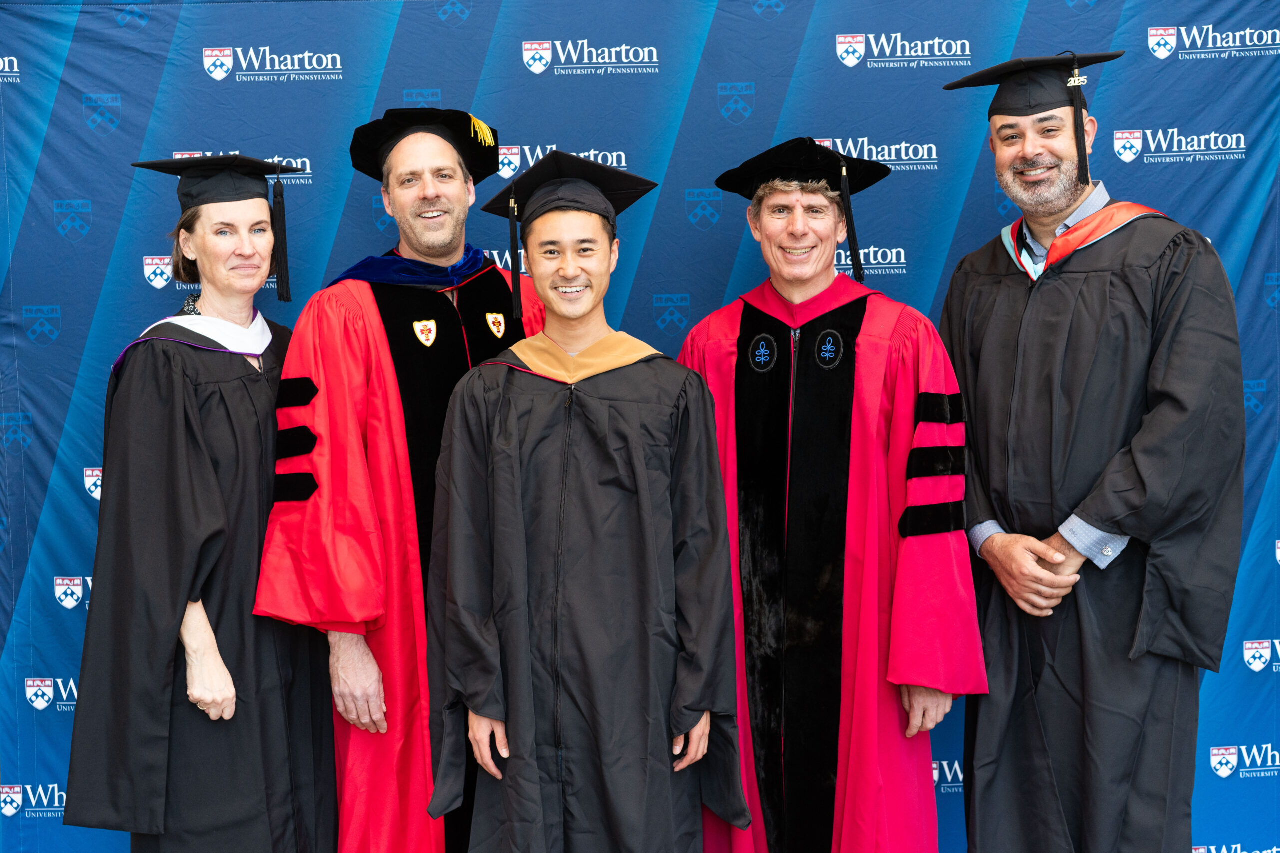 A group of people in academic regalia, featuring several individuals wearing black gowns and mortarboards, and others in red doctoral robes. They are standing in front of a Wharton School of the University of Pennsylvania backdrop, likely at a graduation event.