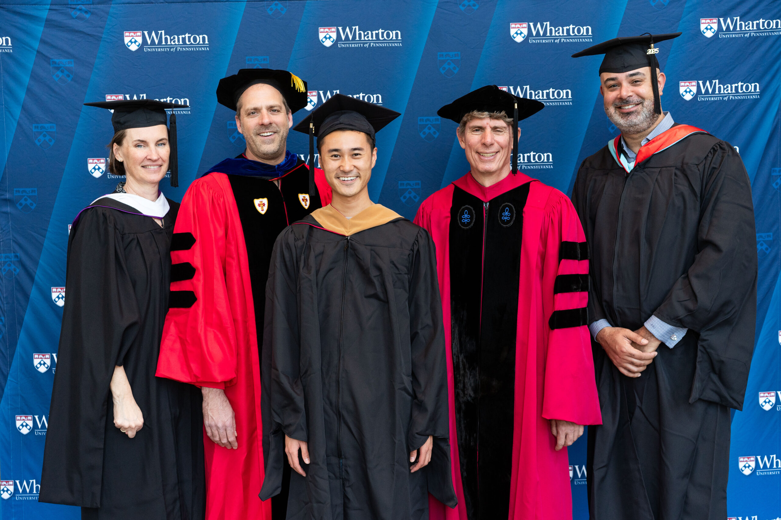 A small group of people wearing academic regalia, including caps and gowns, pose in front of a backdrop with "Wharton University of Pennsylvania" logos, suggesting a graduation or academic ceremony.
