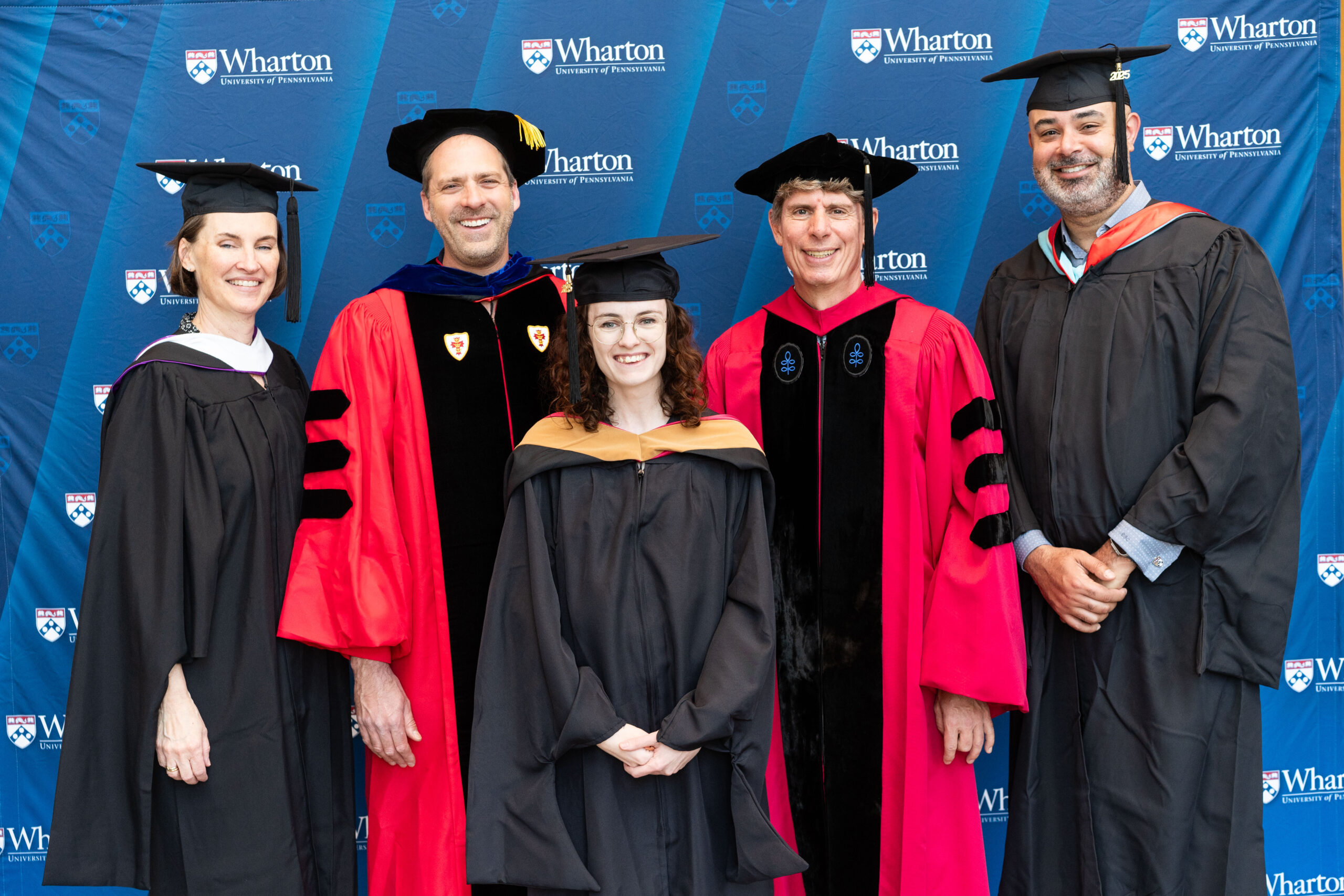 A group of people in academic regalia, including robes and caps, pose against a backdrop with the Wharton School logo indicating a graduation ceremony or academic event.