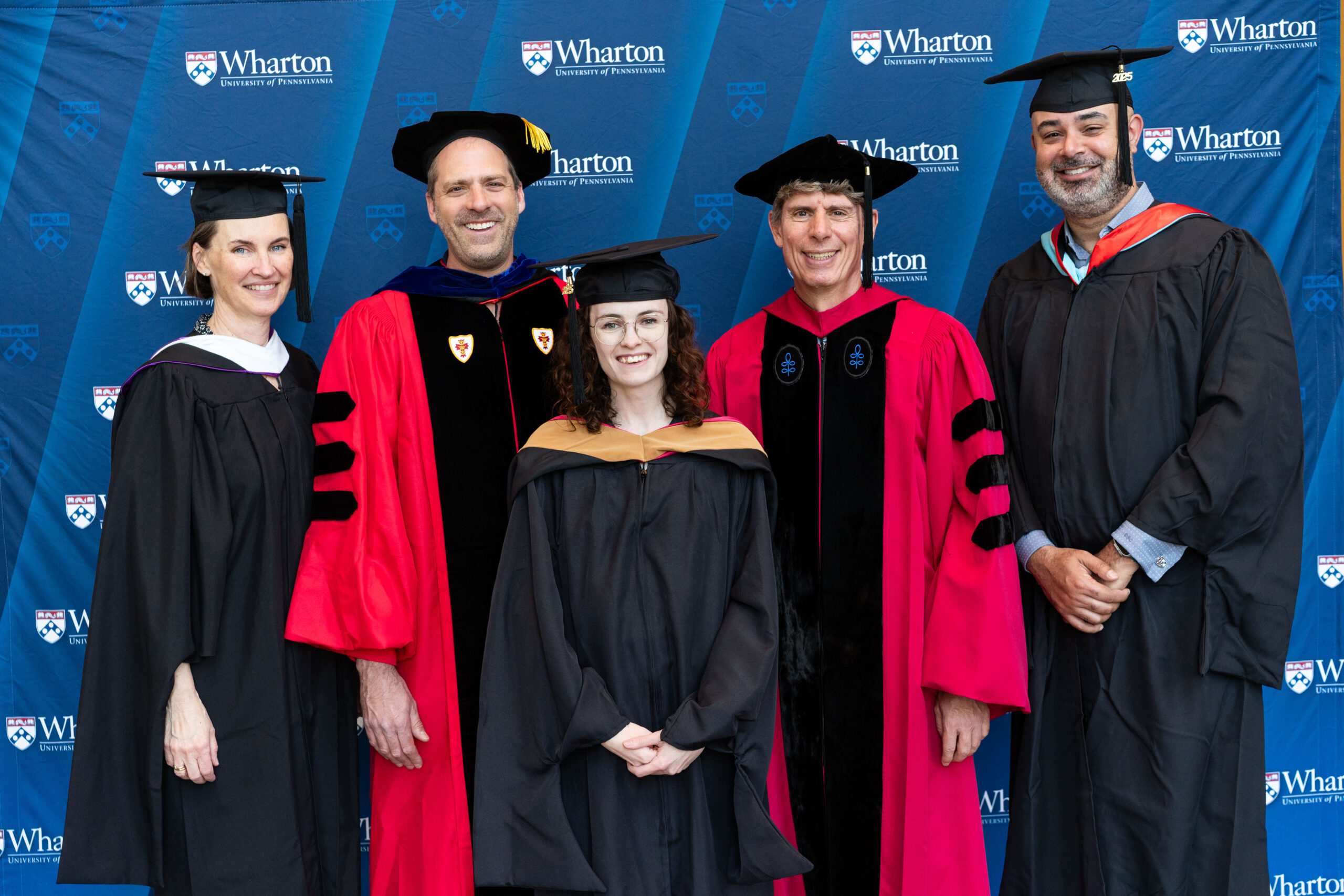 A small group of people in academic regalia, including caps and gowns, smiling in front of a Wharton School backdrop.