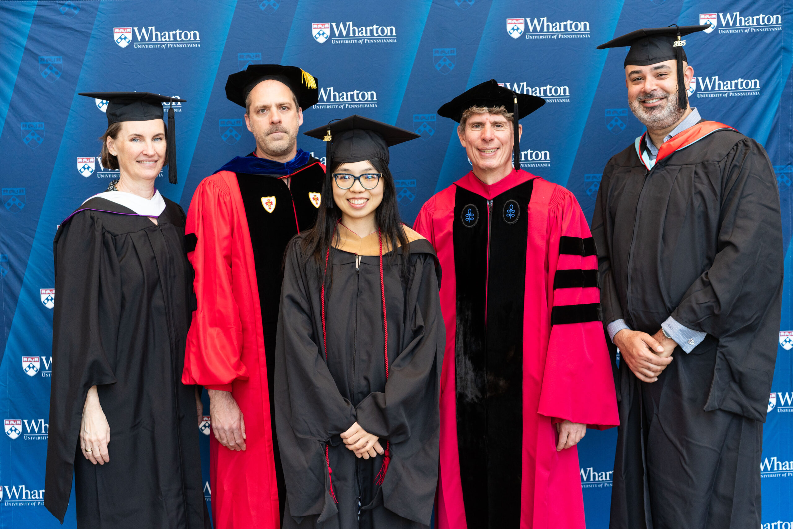 A small group of people in graduation attire pose in front of a backdrop with the Wharton School logo.