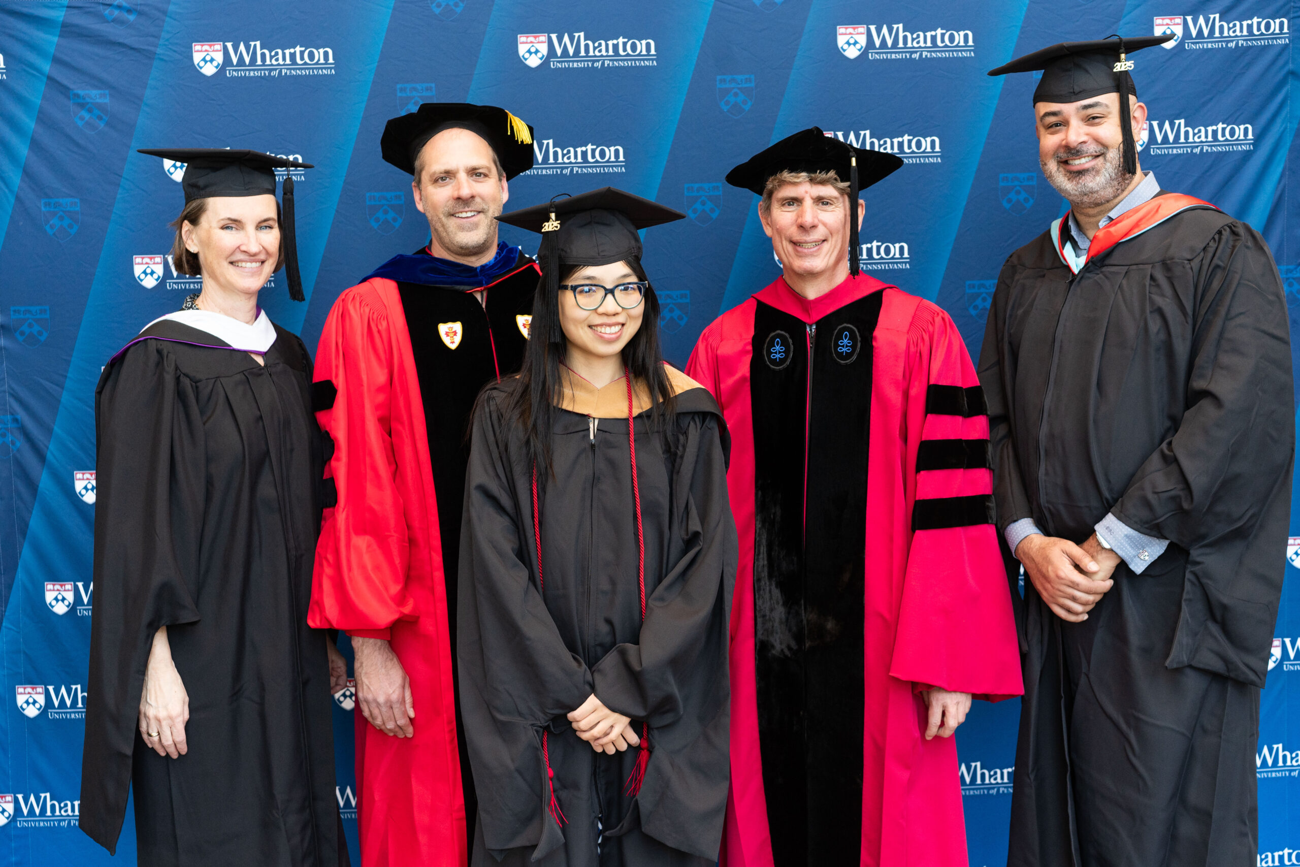 A small group of people in academic caps and gowns, posing in front of a "Wharton University of Pennsylvania" backdrop, suggesting a graduation event.