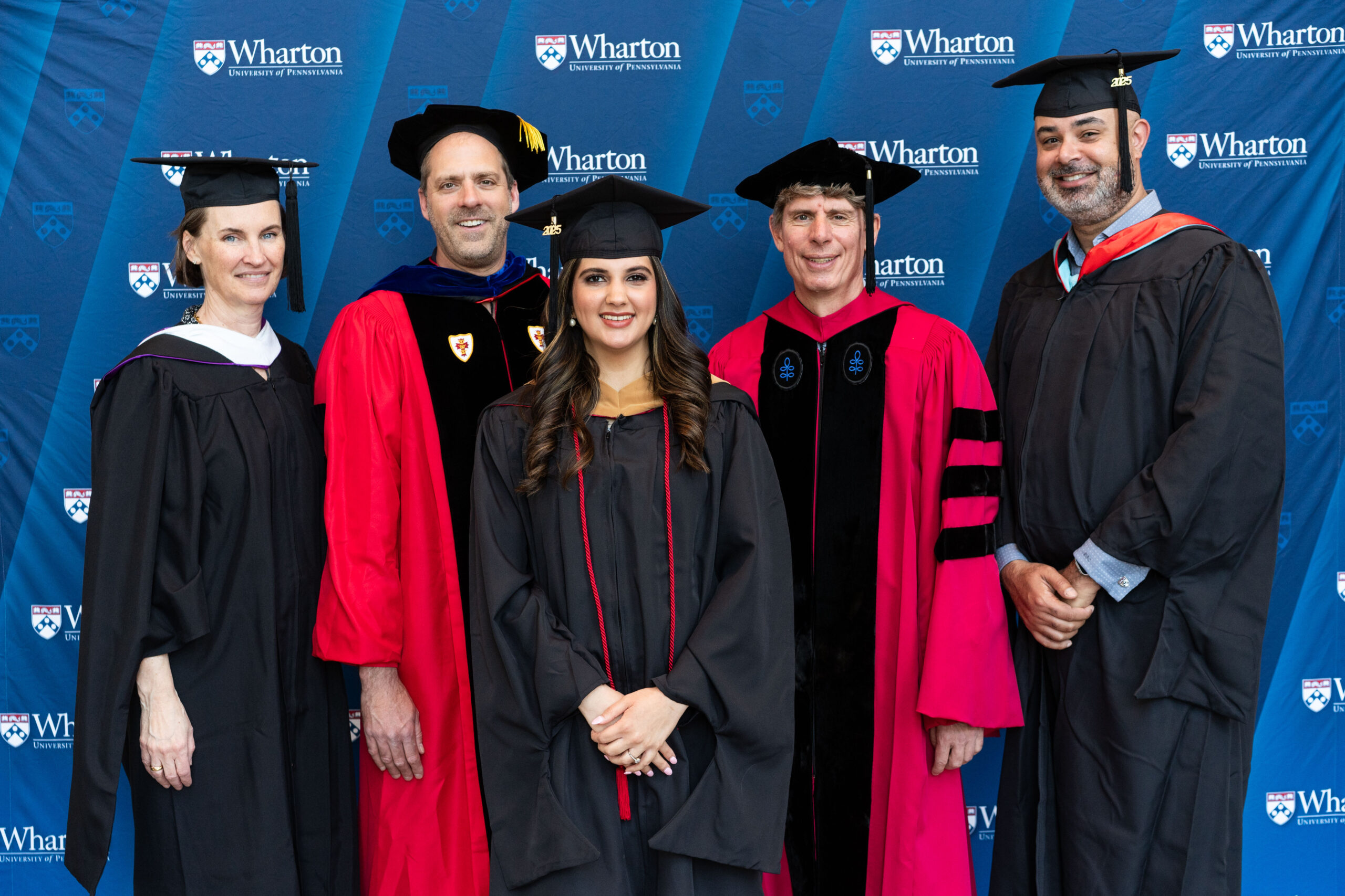 A group of people in graduation and academic regalia, standing in front of a Wharton School University of Pennsylvania backdrop.