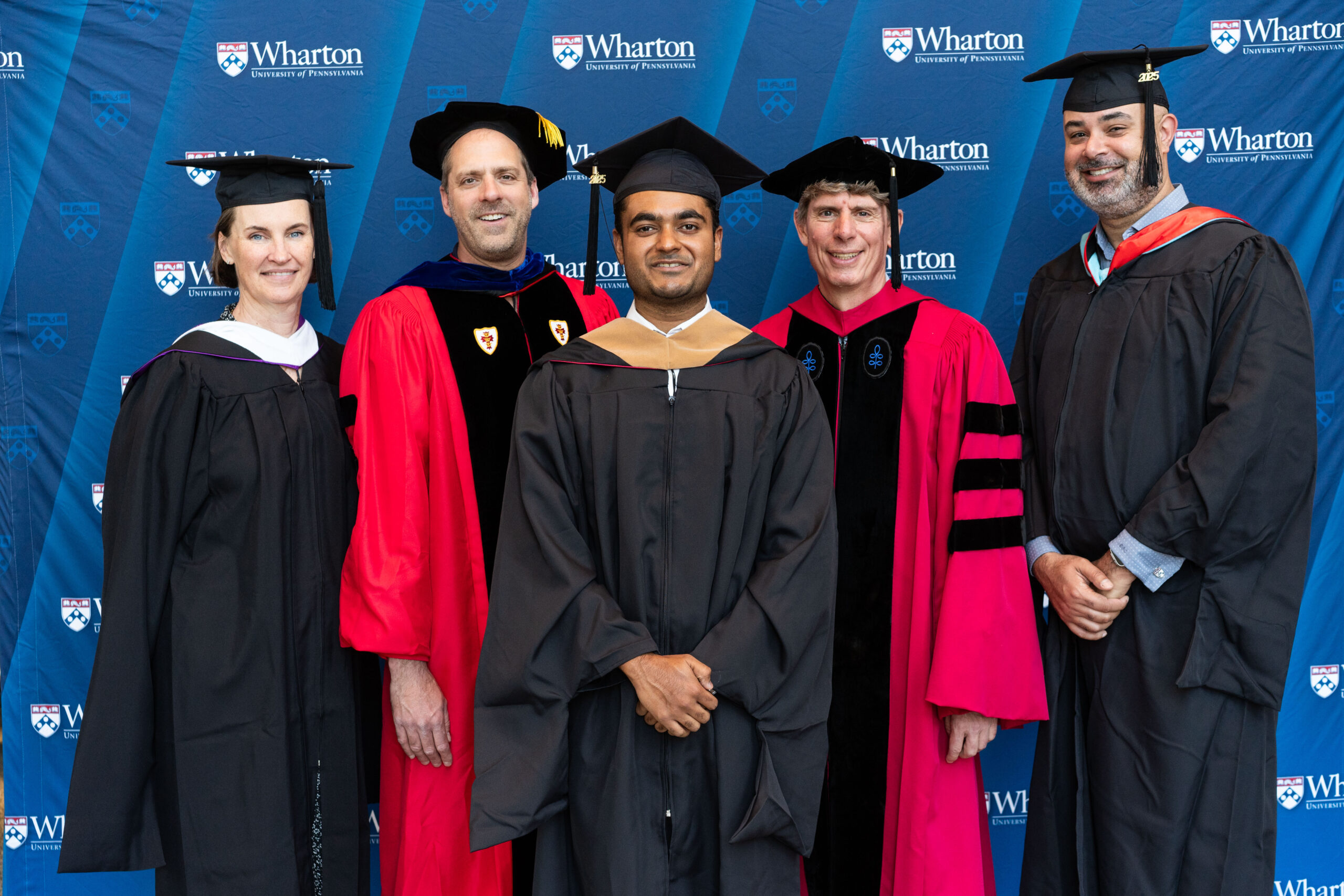 A small group of individuals in academic regalia posing in front of a Wharton University of Pennsylvania backdrop, likely at a graduation event.