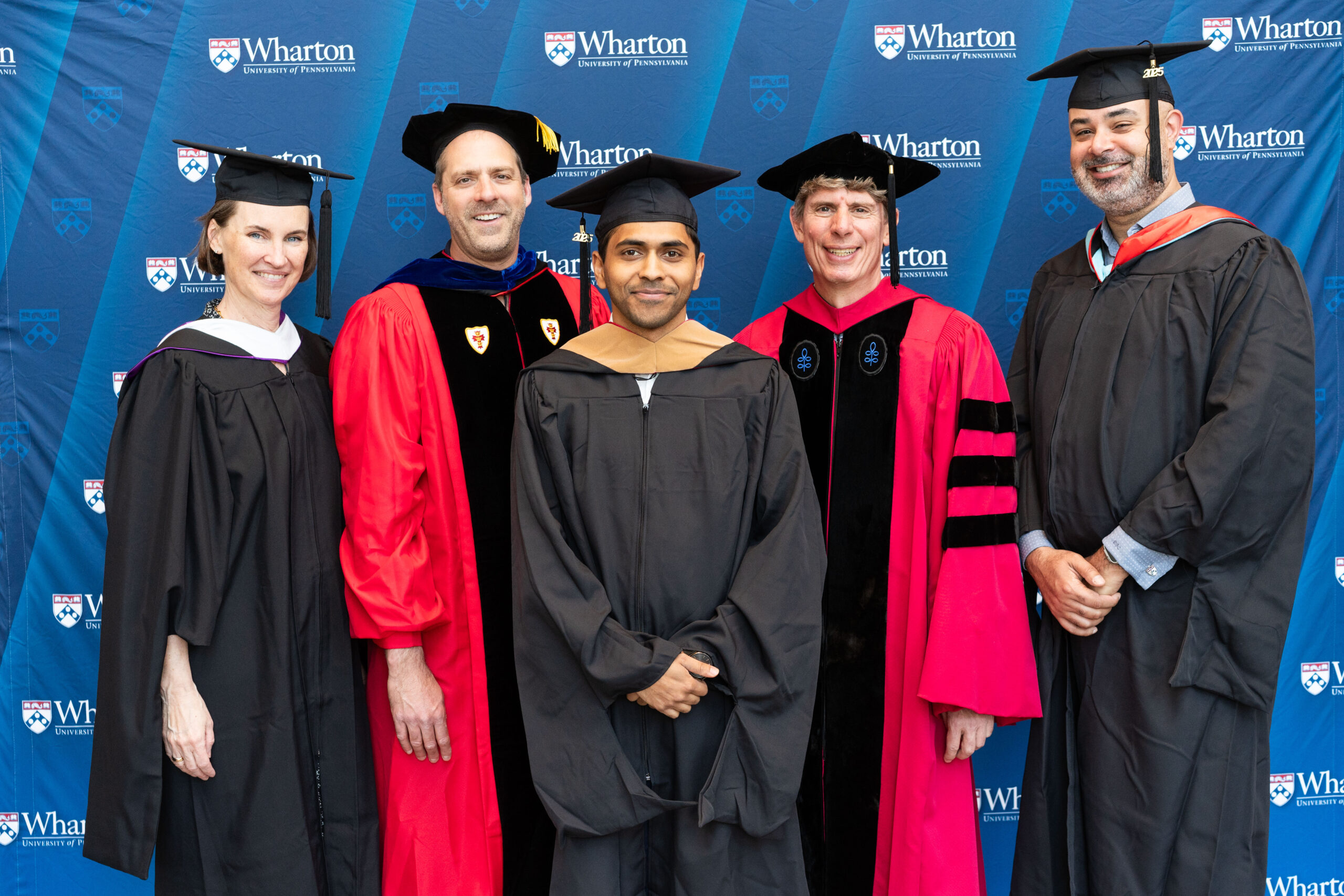 A group of people in academic regalia posing for a graduation photo against a backdrop that reads "Wharton University of Pennsylvania."