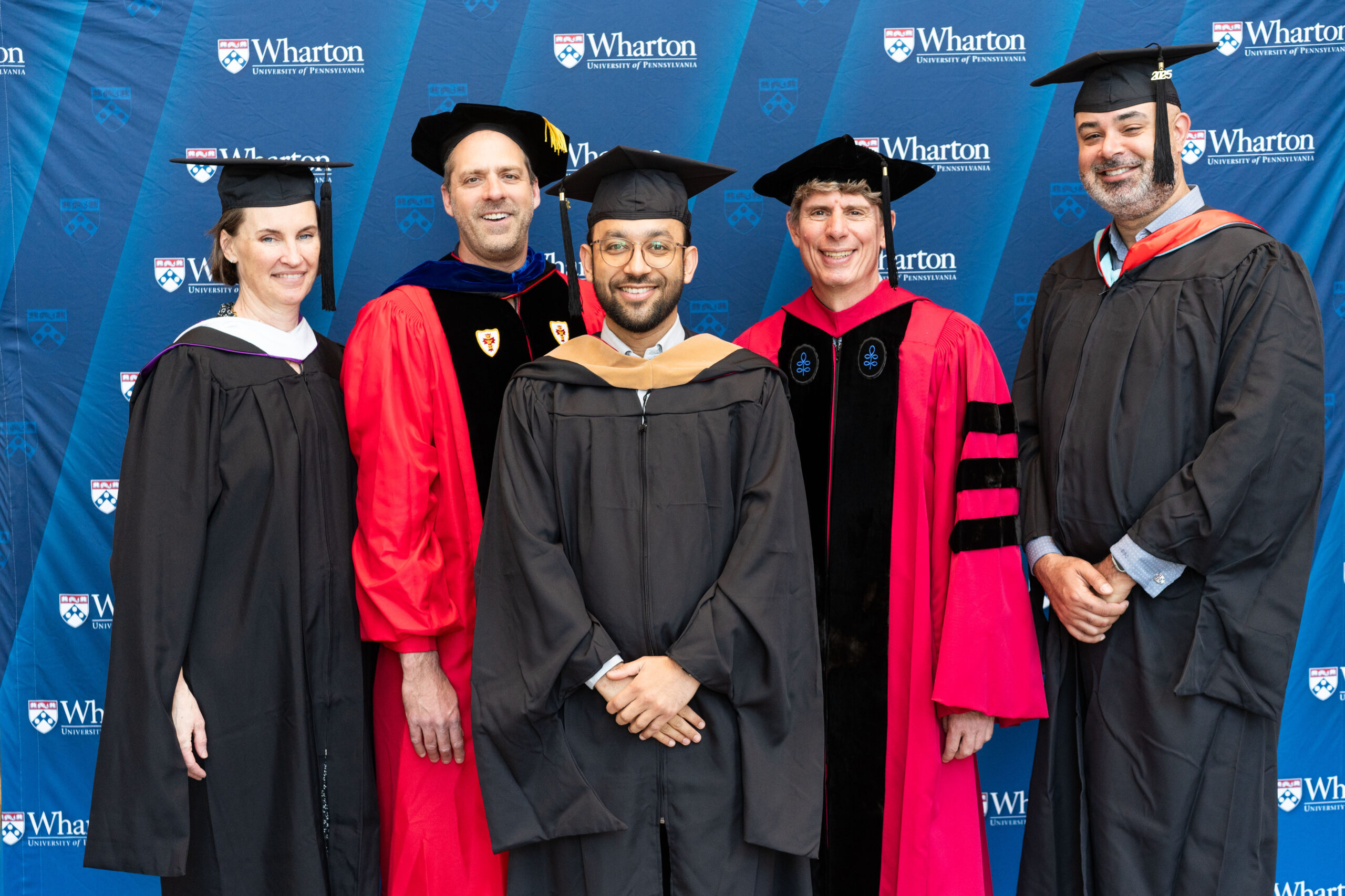 A small group in academic regalia, including graduates and faculty, posing in front of a Wharton School backdrop.