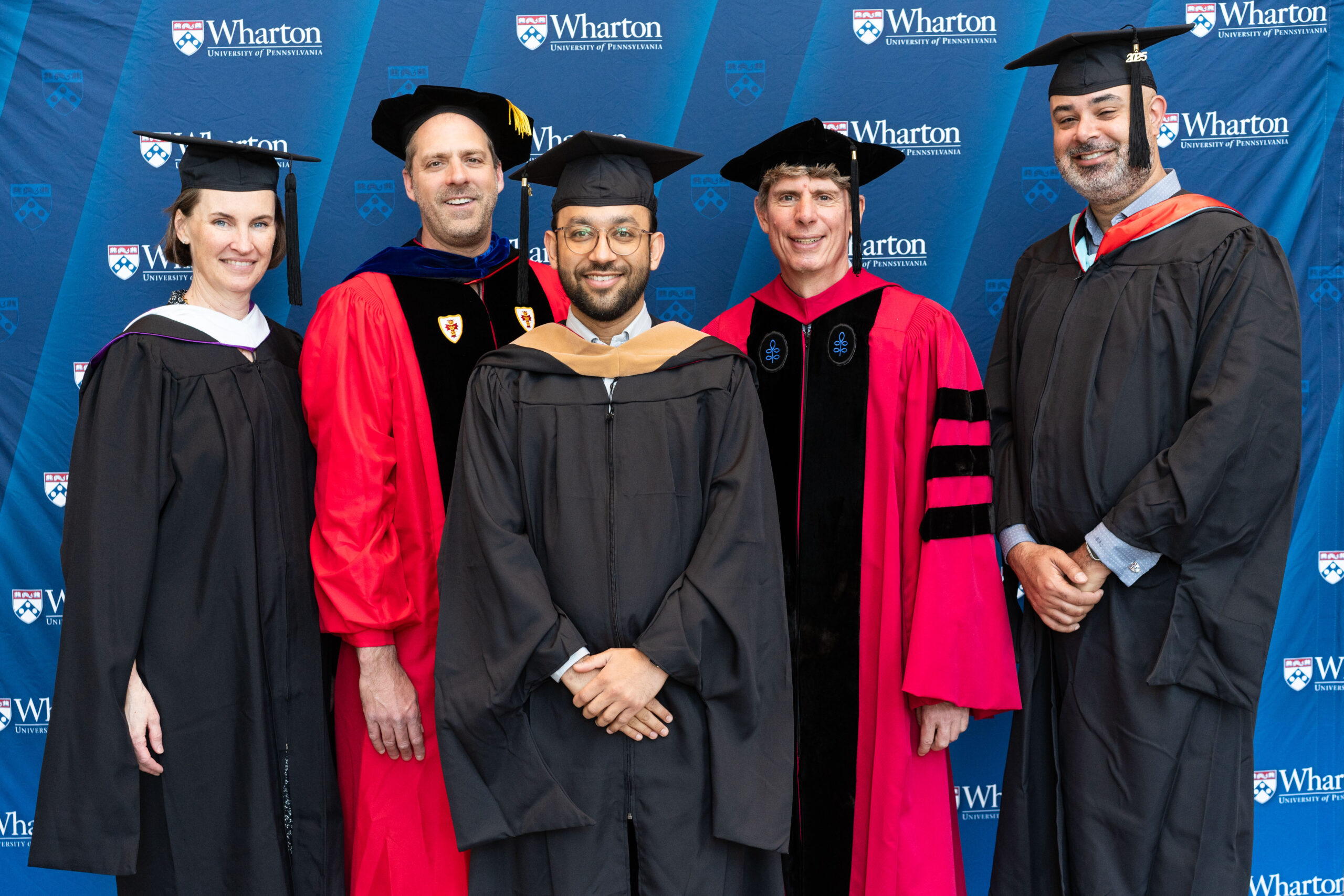 A group of people in academic regalia, including caps and gowns, standing in front of a backdrop with the Wharton School of the University of Pennsylvania logo, possibly celebrating a graduation event.