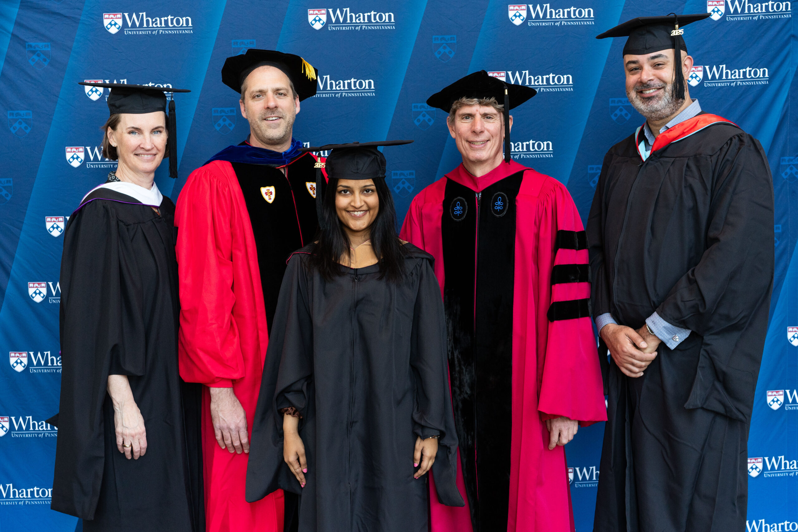 A small group of graduates in caps and gowns, posing in front of a "Wharton University of Pennsylvania" backdrop.