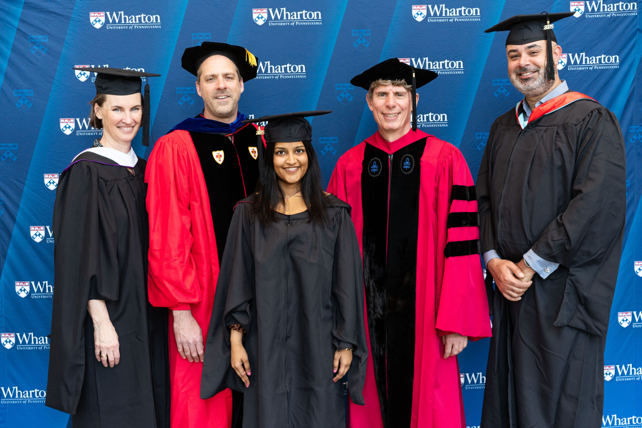 A small group of individuals in academic regalia, standing in front of a Wharton School backdrop, likely representing a graduation or academic event.