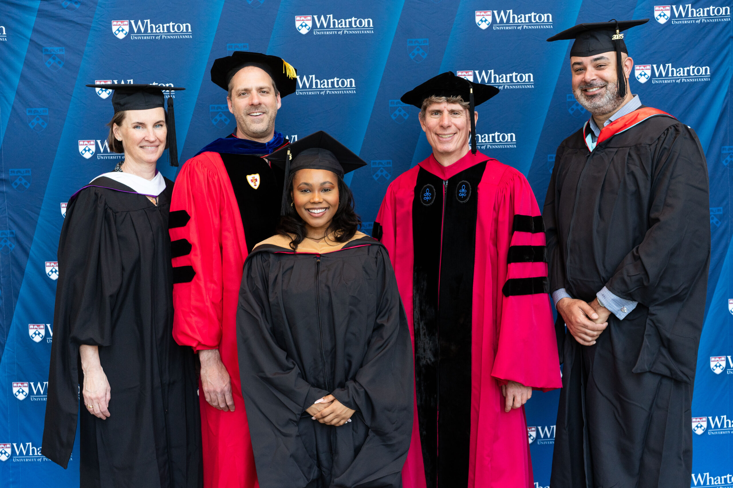 A group of people in graduation attire, including robes and caps, pose for a photo in front of a backdrop featuring the Wharton School logo.