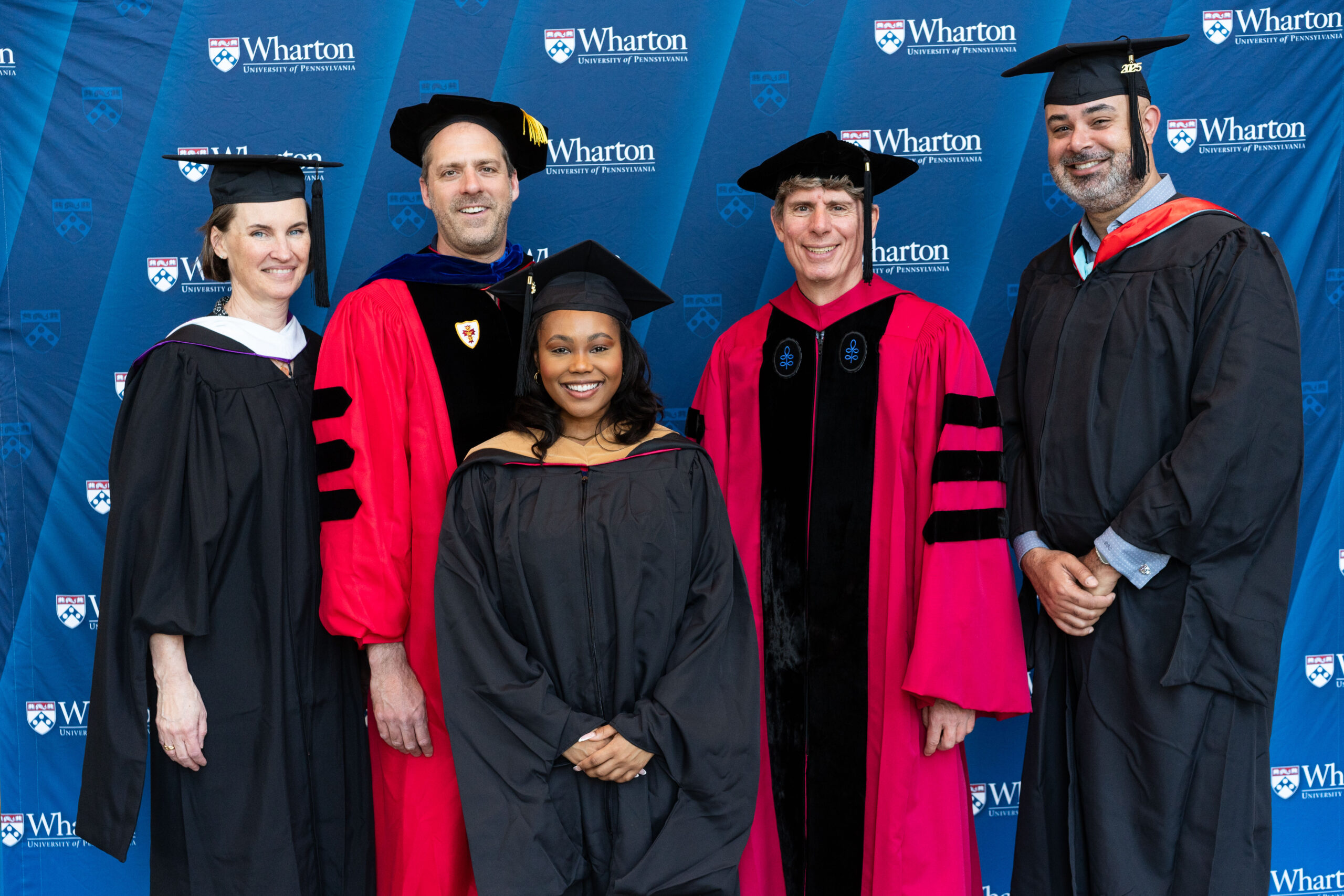 A group of five individuals in academic regalia, including caps and gowns, stand smiling in front of a Wharton School of the University of Pennsylvania backdrop.