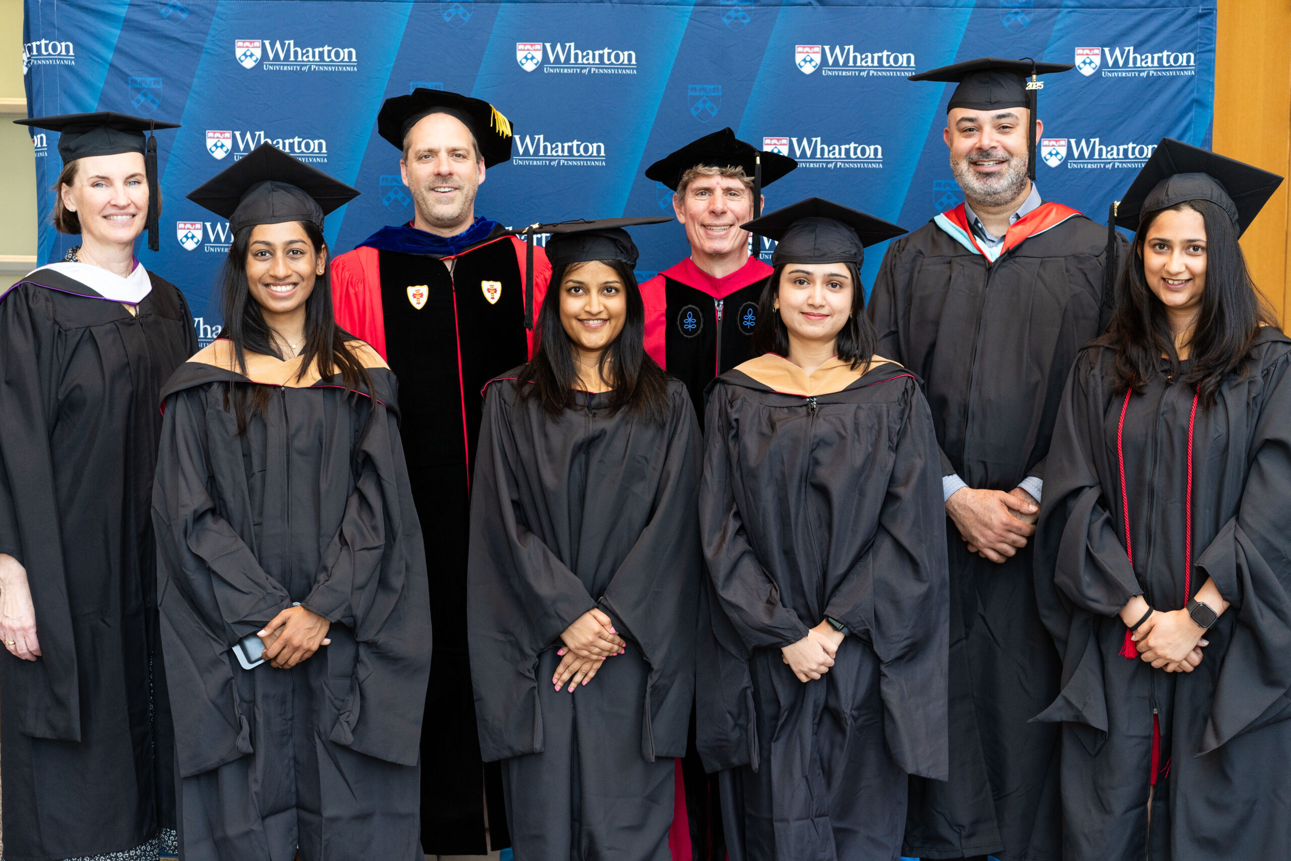 A small group of graduates and faculty members in academic regalia are posing for a photo in front of a Wharton School banner.
