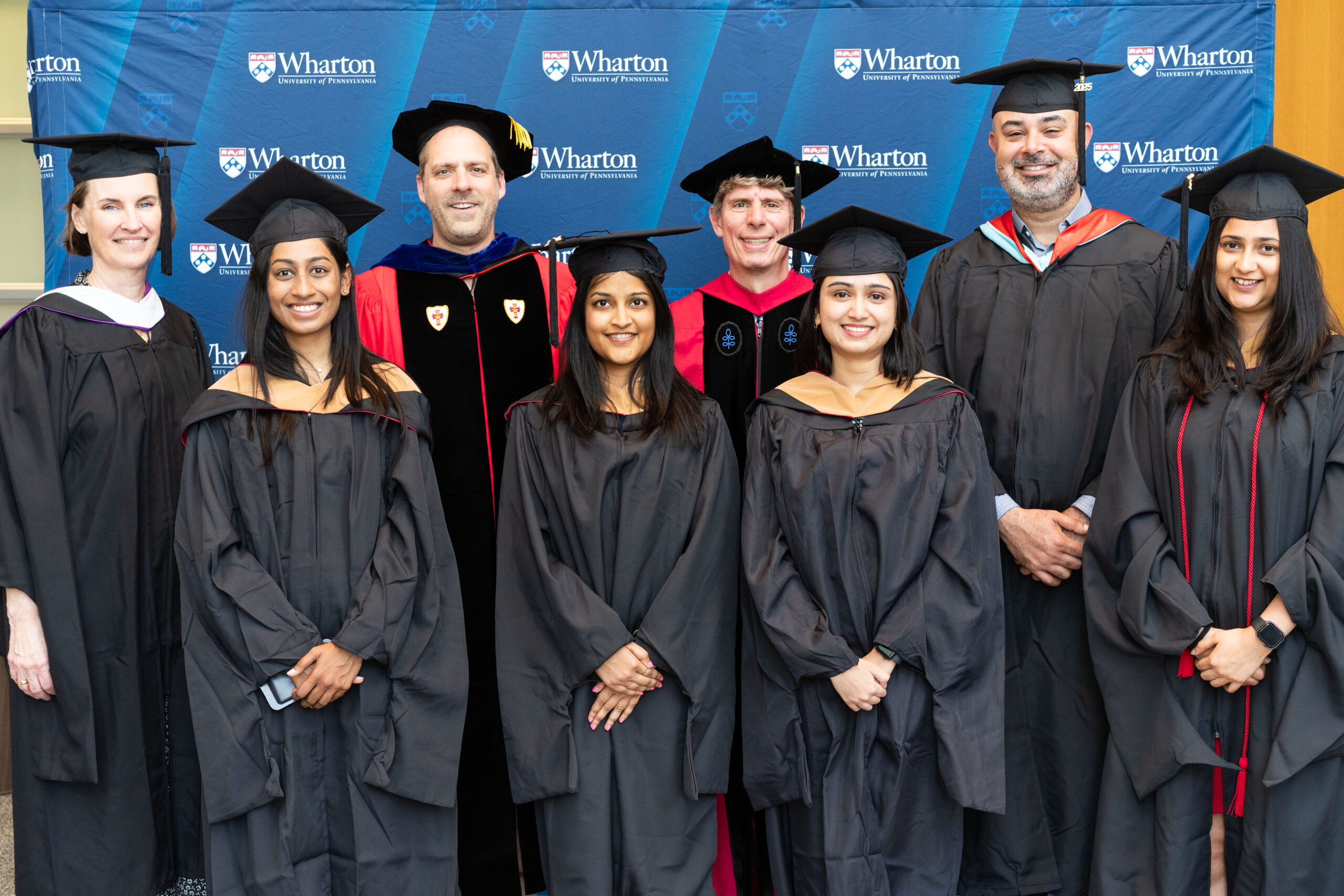 A group of graduates in caps and gowns stands in front of a backdrop with the Wharton School logo, alongside faculty members in academic regalia.